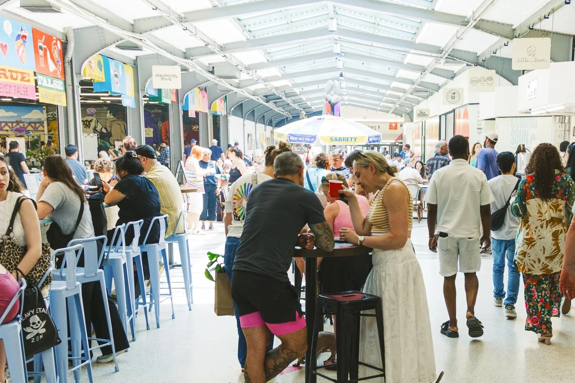 A crowded indoor marketplace or food court with people sitting and standing, some using laptops and phones. There are high chairs, tables, and a large umbrella with advertising. Bright lighting and signs are visible overhead.
