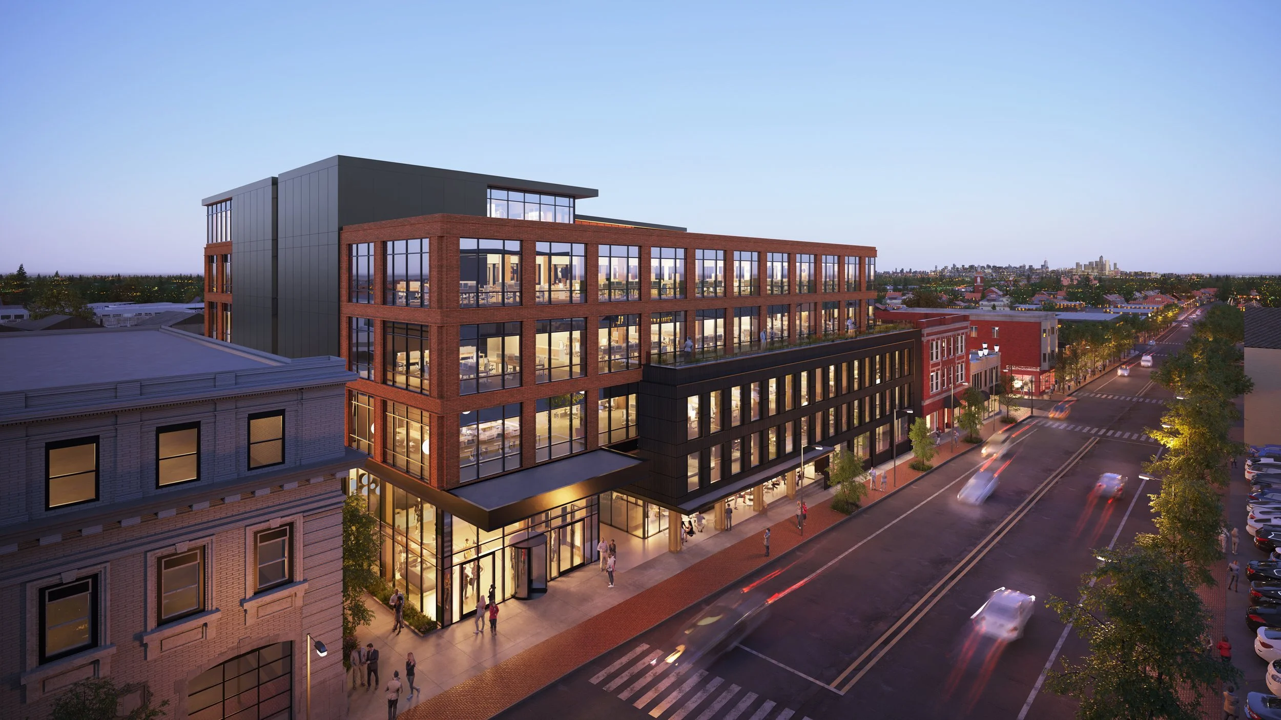 A multi-story modern building with large glass windows, situated in an urban area during dusk. The building features a red brick facade and a dark upper section, with people walking on the sidewalk and cars on the street.