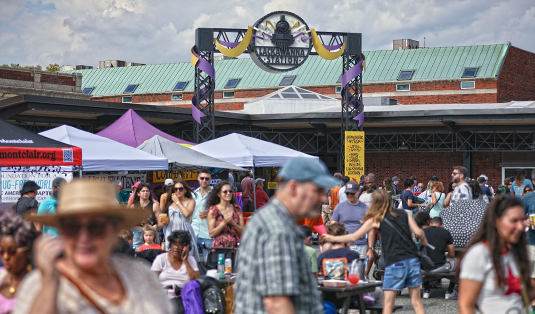 Crowd gathered at an outdoor event in front of LACKAWANNA STATION, with tents and booths, some people sitting and others standing, under partly cloudy skies.