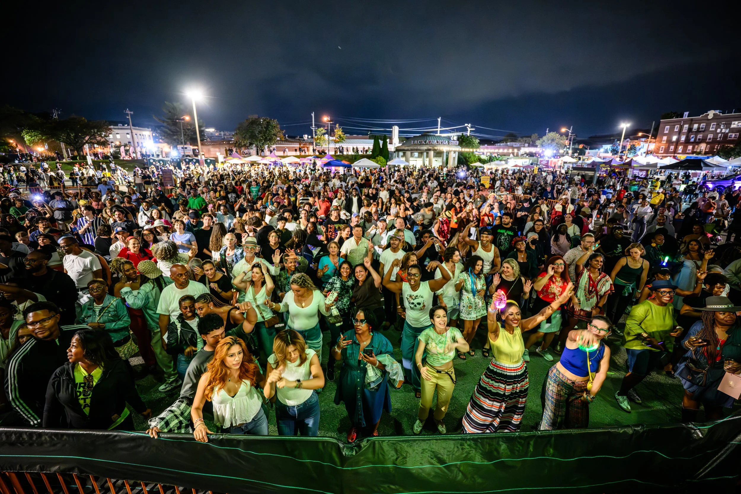 Crowd of people at an outdoor event at night, enjoying music, dancing, and socializing with tents and lights in the background.
