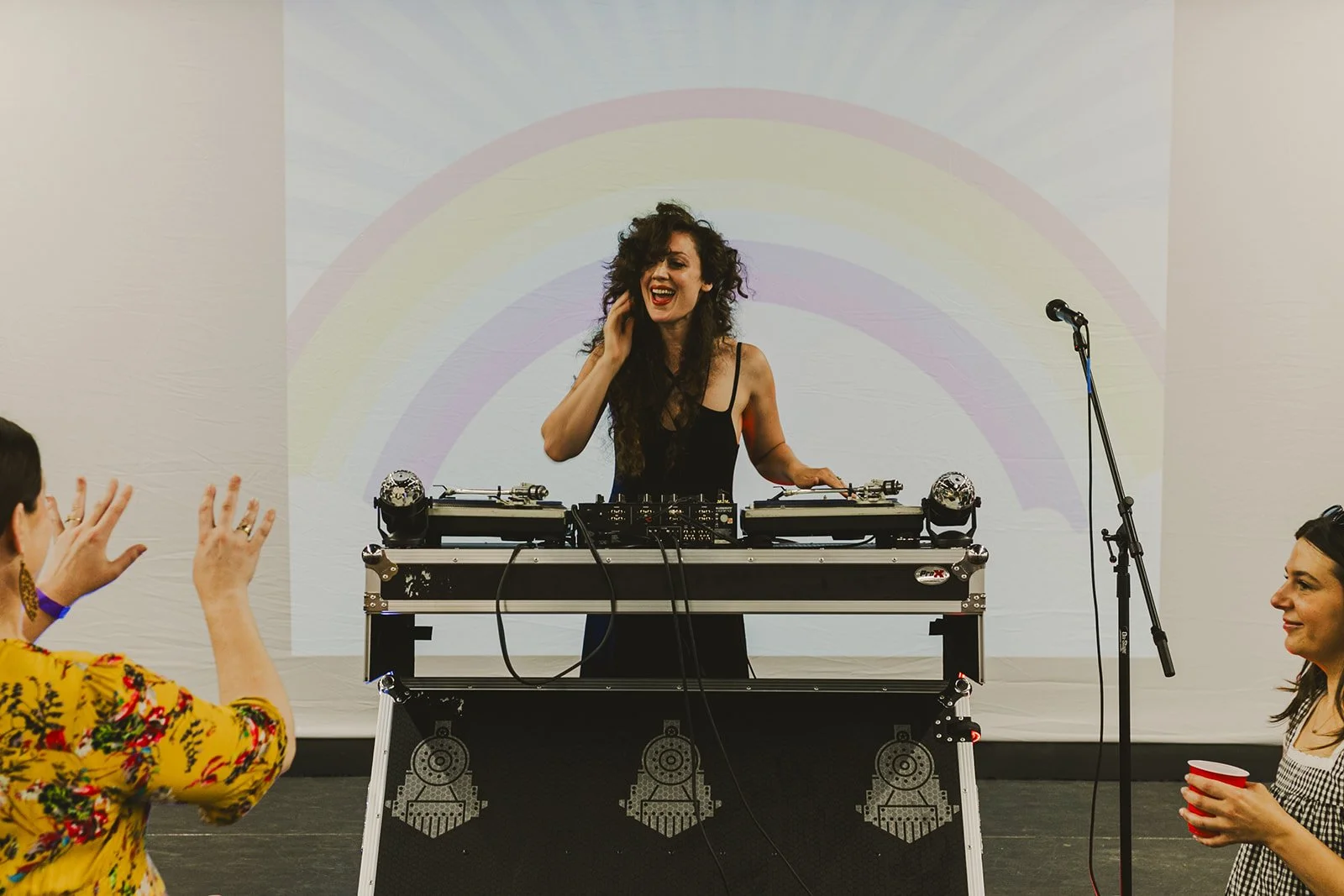 A woman DJ with curly hair performing at a party, smiling, with people dancing and enjoying in front of her, against a backdrop with a rainbow projection, microphone stand on the right, and a white wall.