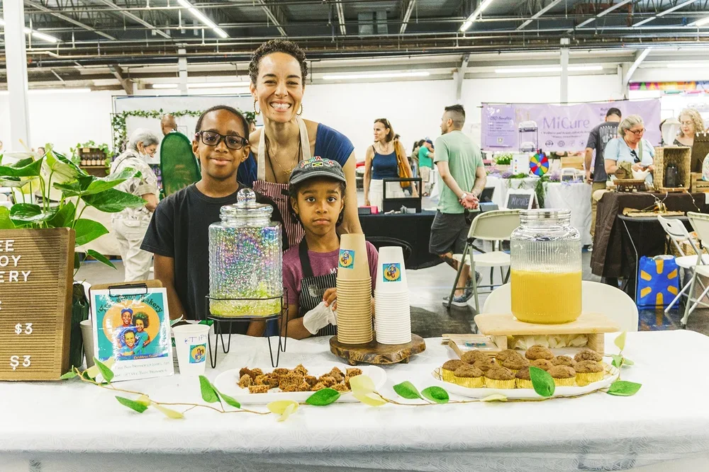 Three people, two children and an adult woman, standing behind a table with lemonade and baked goods at an indoor market. The table has paper cups, a large jar of lemonade, a plate of cookies, and a decorative sign. The background shows other market stalls and people walking around.