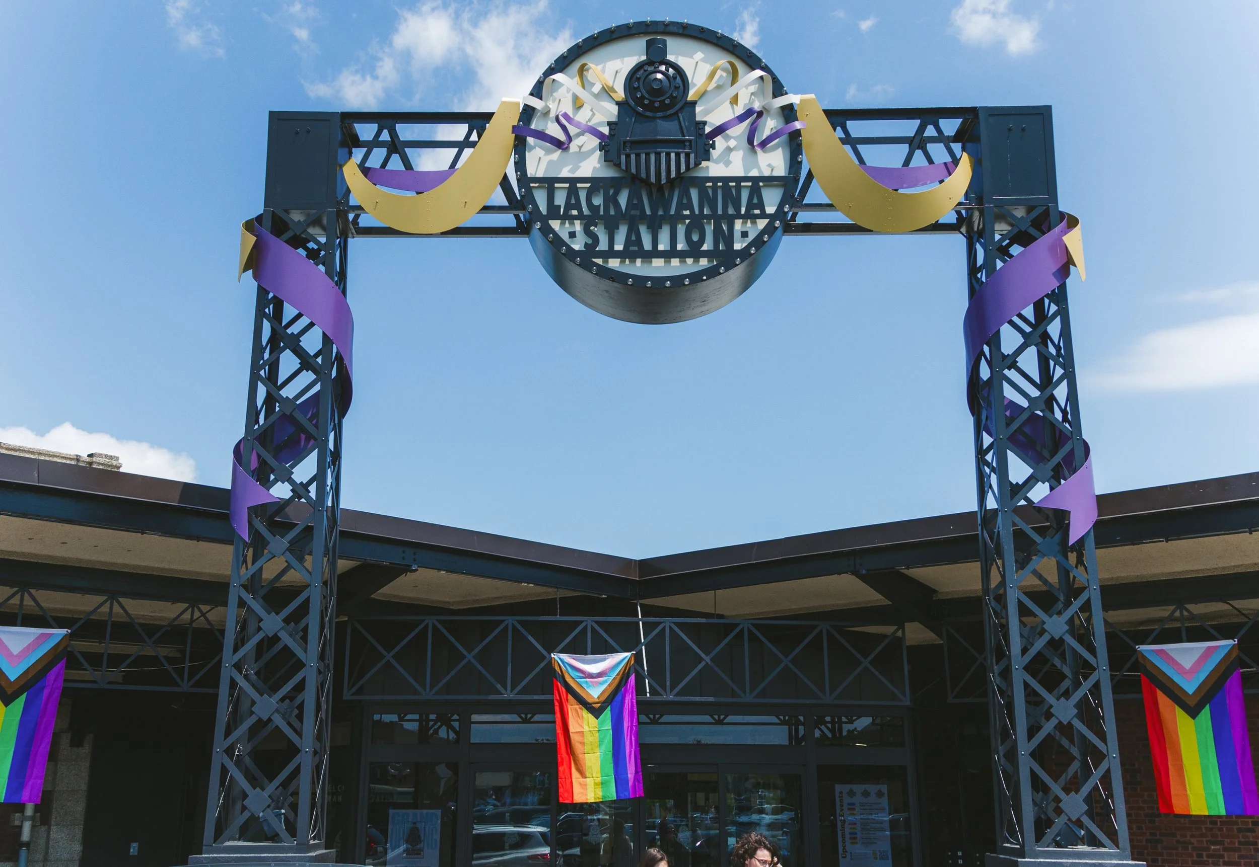 The entrance of a train station with a prominent sign reading 'Lackawanna Station,' decorated with rainbow flags and bunting, against a bright blue sky with some clouds.