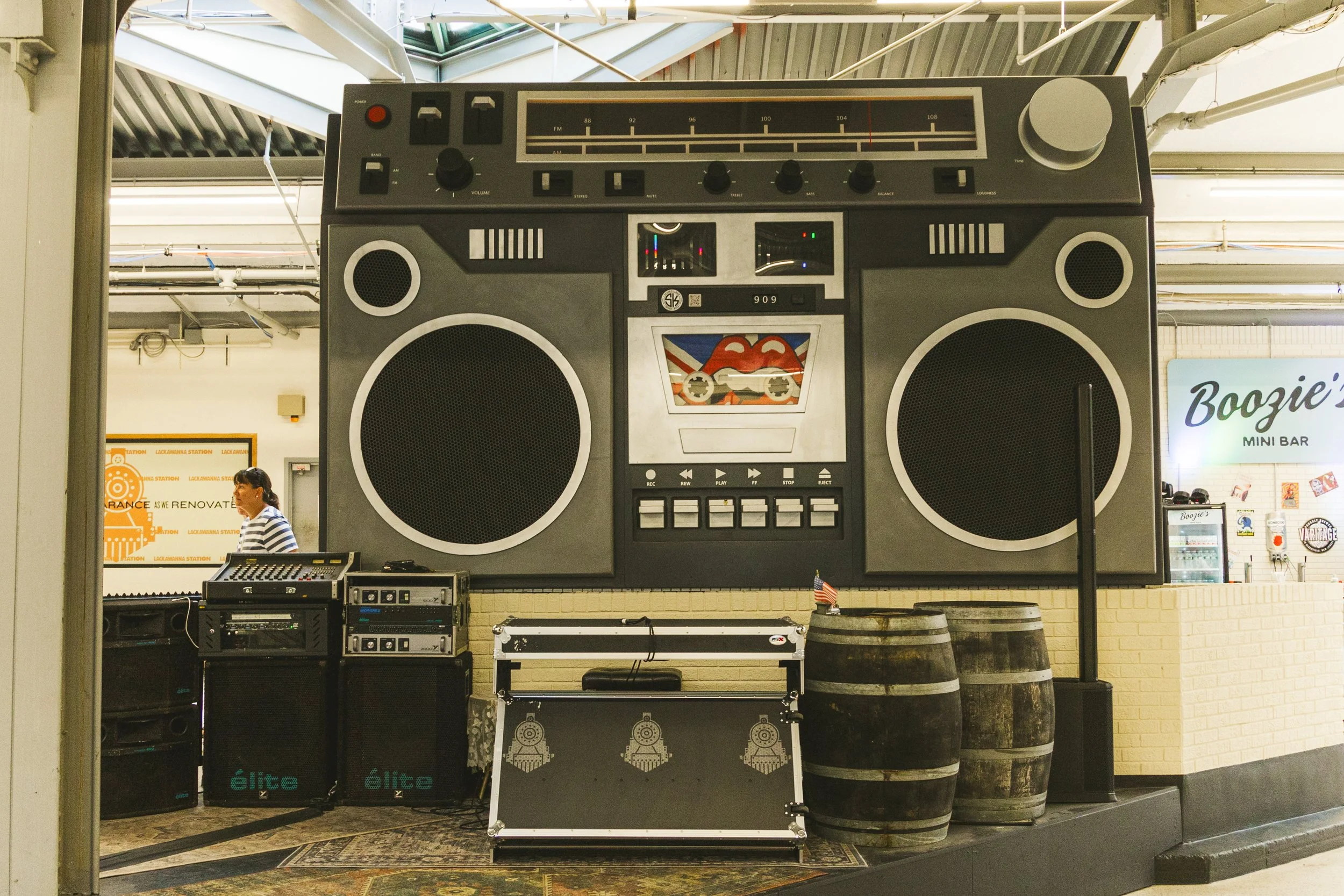 Large vintage Boombox speaker system with a radio tuner, cassette player, and speakers, set in an indoor public space with industrial ceiling and a person in the background.