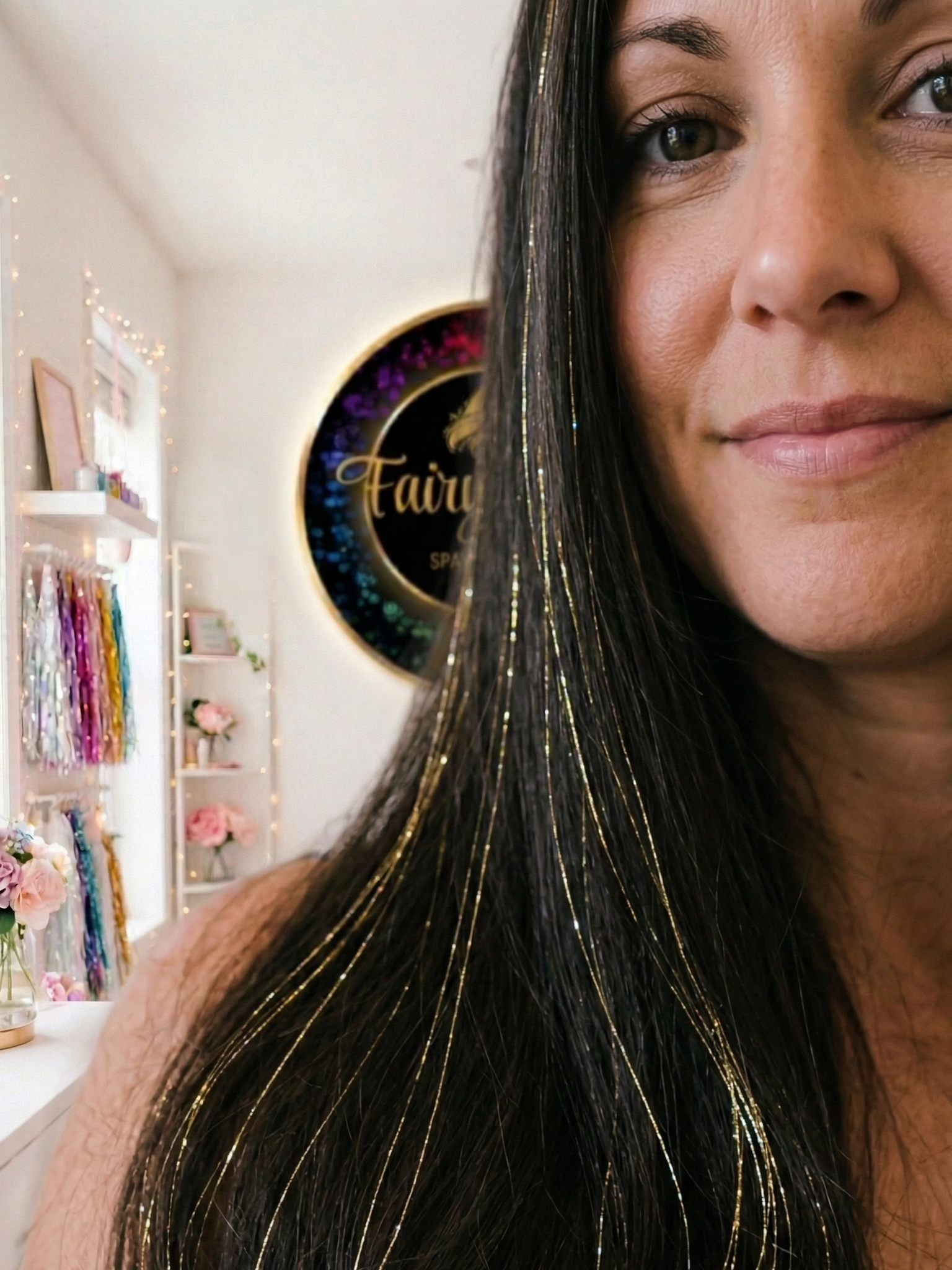Close-up of a woman with long black hair with fairy / tinsel hair, smiling softly, inside a decorated space with a black and gold sign reading 'Fairy' in the background and shelves with flowers and colorful items.