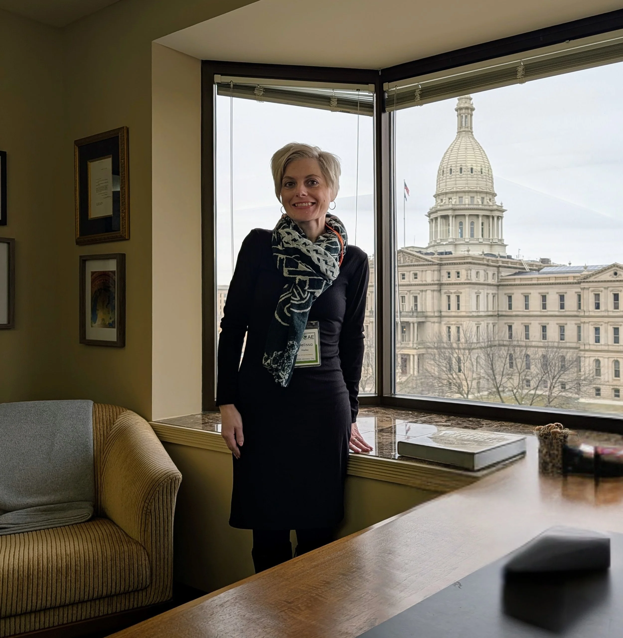 A woman with short blonde hair and a black dress standing inside an office near large windows, with the United States Capitol building visible in the background.
