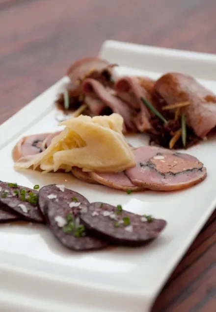 A white rectangular plate with three types of sliced cured meats, including dark sausage slices garnished with chopped green herbs, pale yellow slices of ham, and rolled slices of beef with a sprig of rosemary.