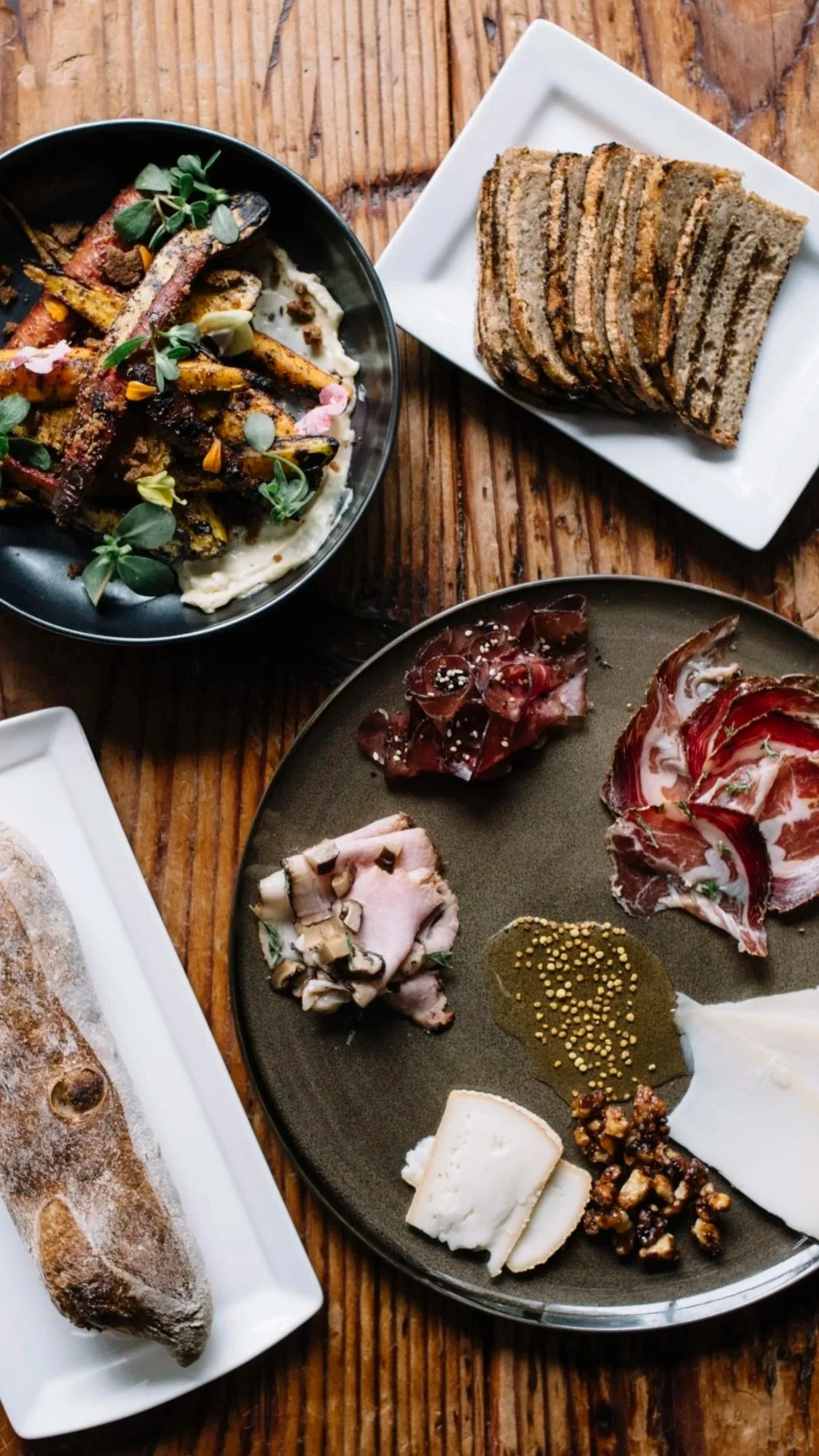 Assorted charcuterie including sliced cheeses, cured meats, and condiments on a black plate, beside a loaf of bread on a white platter, all on a wooden table.