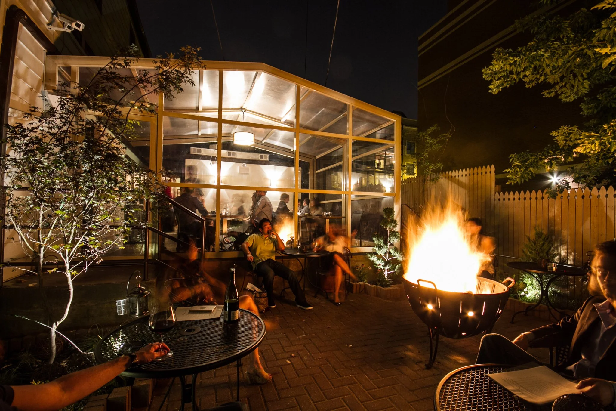 Night scene of a backyard gathering with a glass-enclosed patio, outdoor fire pit, and several people sitting around, illuminated by the fire and patio lights, with trees and a wooden fence.