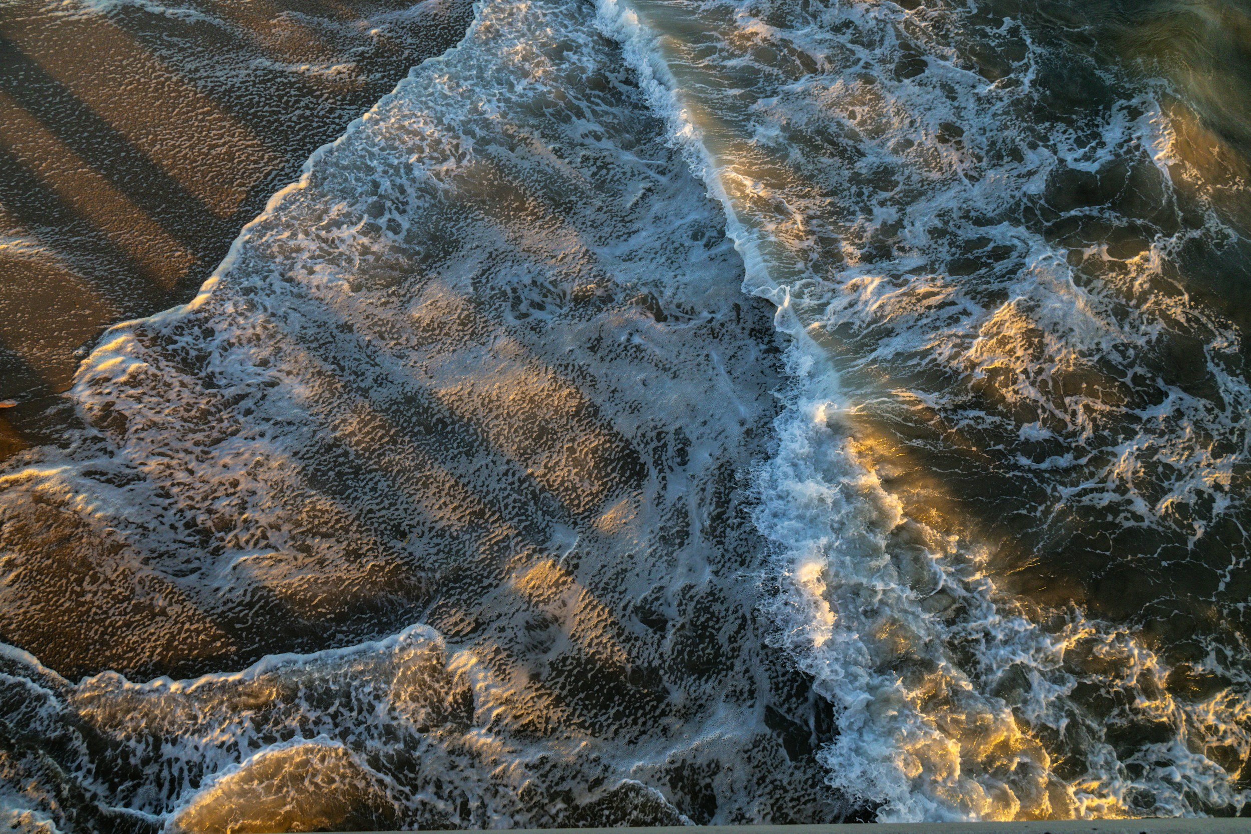 Aerial view of ocean waves crashing onto sandy beach during sunset.