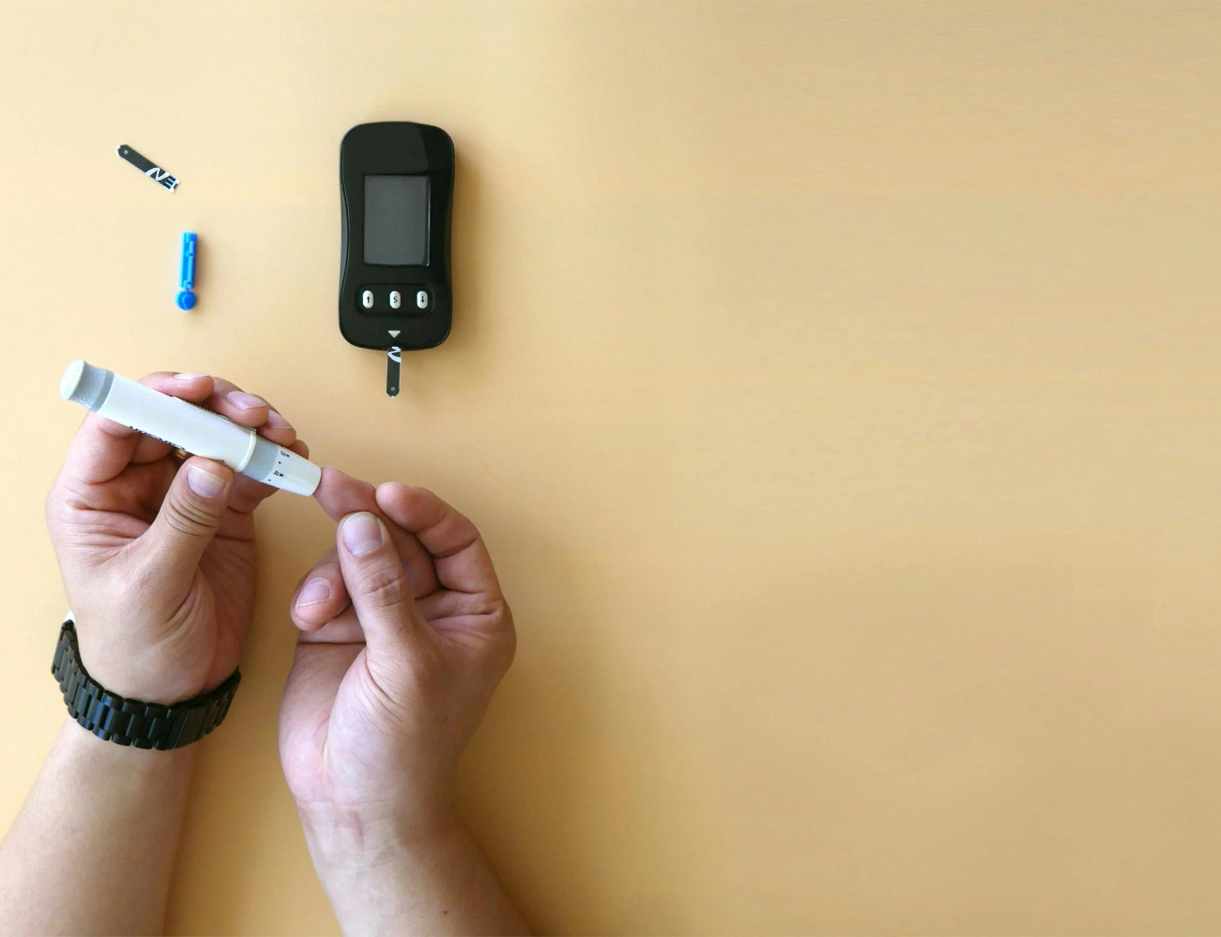 Person holding a white marker pen near a digital glucometer on a yellow surface, with a small blue and black adhesive bandage and a black watch on their wrist.