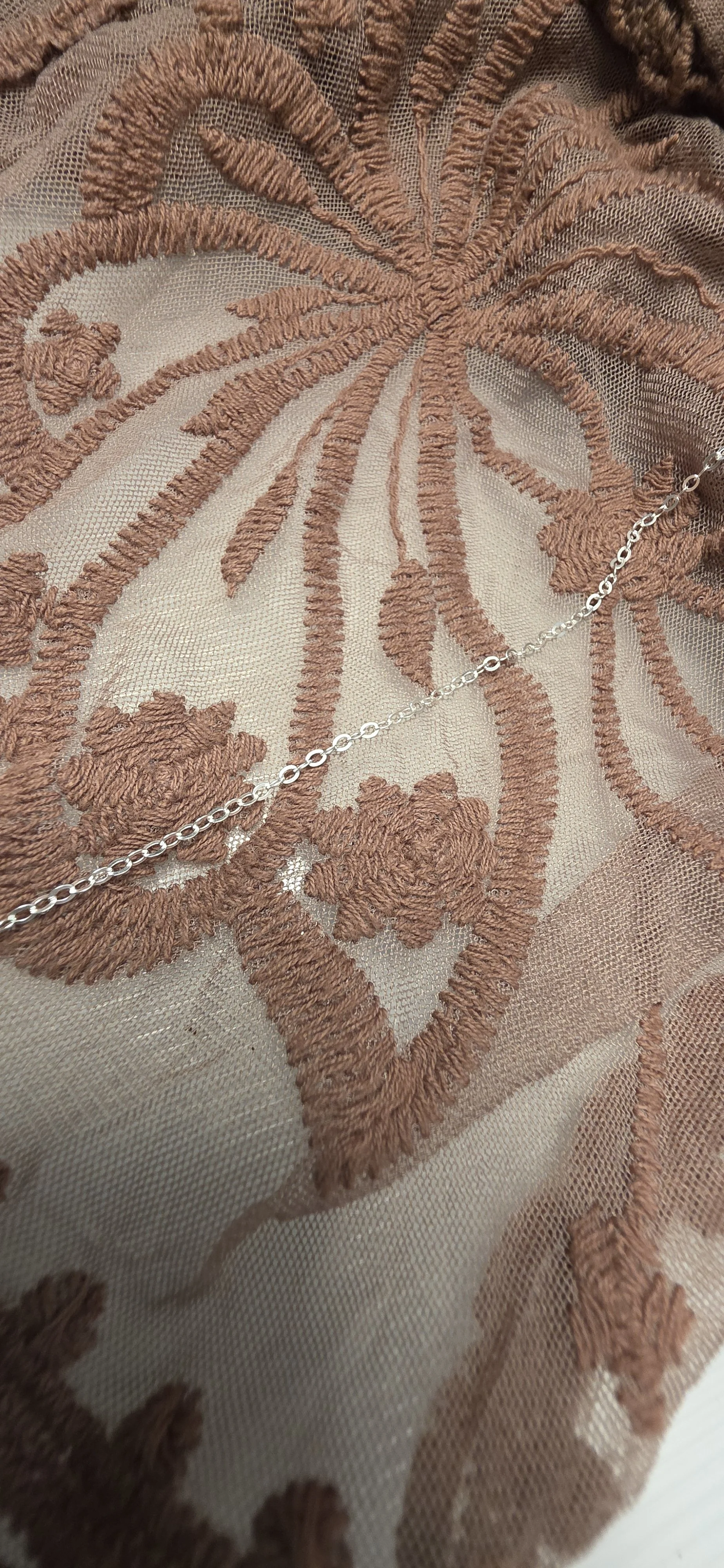 A silver chain necklace resting on embroidered beige fabric with a floral and leaf pattern.