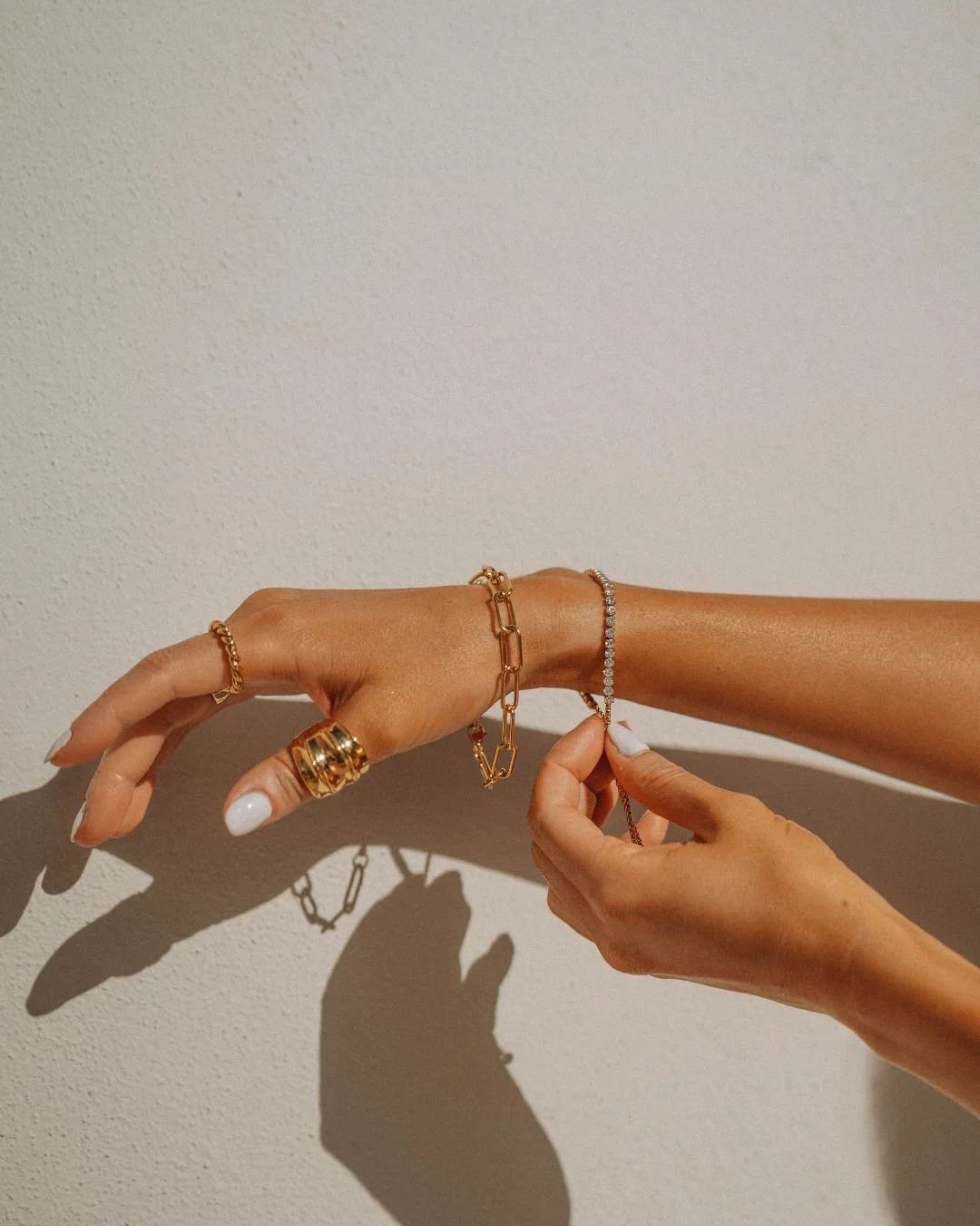 Close-up of a woman's hand and forearm with several gold and diamond jewelry pieces, including rings, bracelets, and a delicate chain bracelet, casting shadows on a plain wall.