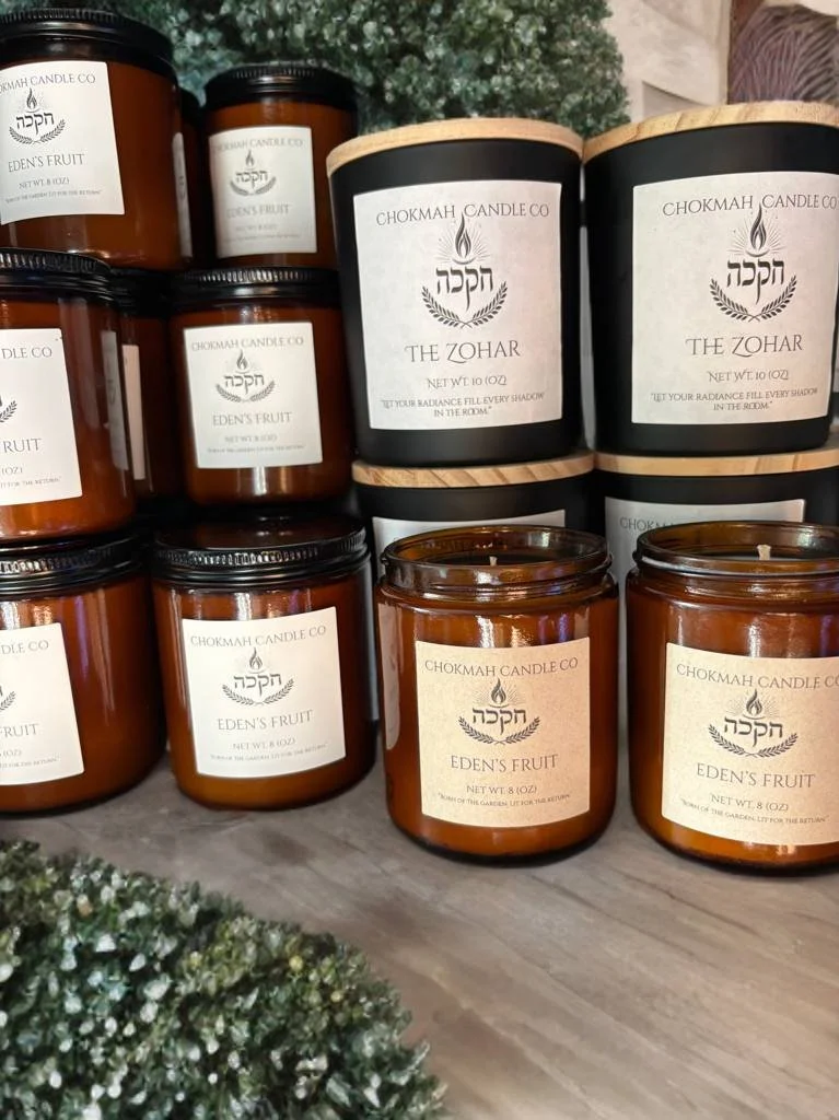 Various candles in amber jars with white labels, including 'Eden's Fruit' and 'The Zohar', displayed on a wooden surface with some greenery in the foreground.