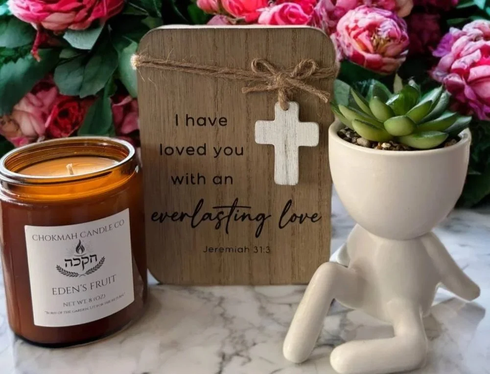 Decorative arrangement with a lit candle labeled 'Eden's Fruit,' a wooden sign with a cross and the message 'I have loved you with an everlasting love,' and a small potted succulent plant in a white pot shaped like a person, all set on a marble surface with a background of pink flowers.