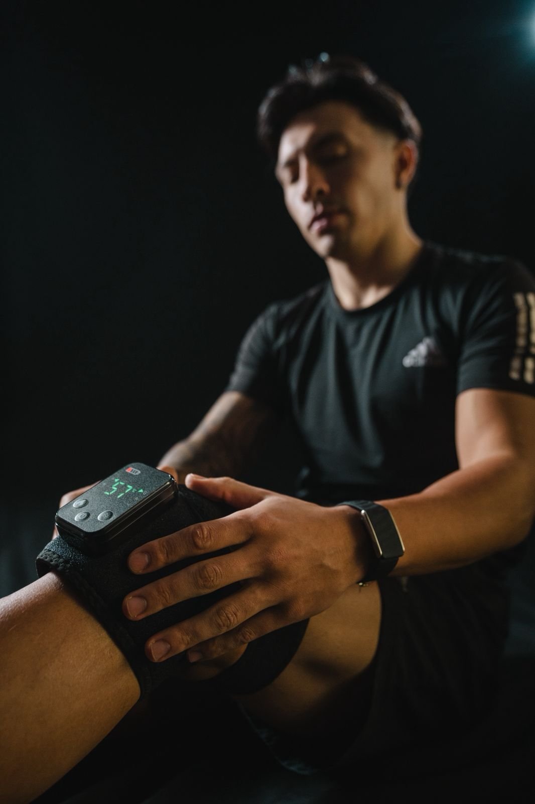 Un hombre joven, con camiseta negra y reloj inteligente, revisa la rodilla de otra persona que lleva una banda con un dispositivo digital, en un entorno oscuro y con poca luz.