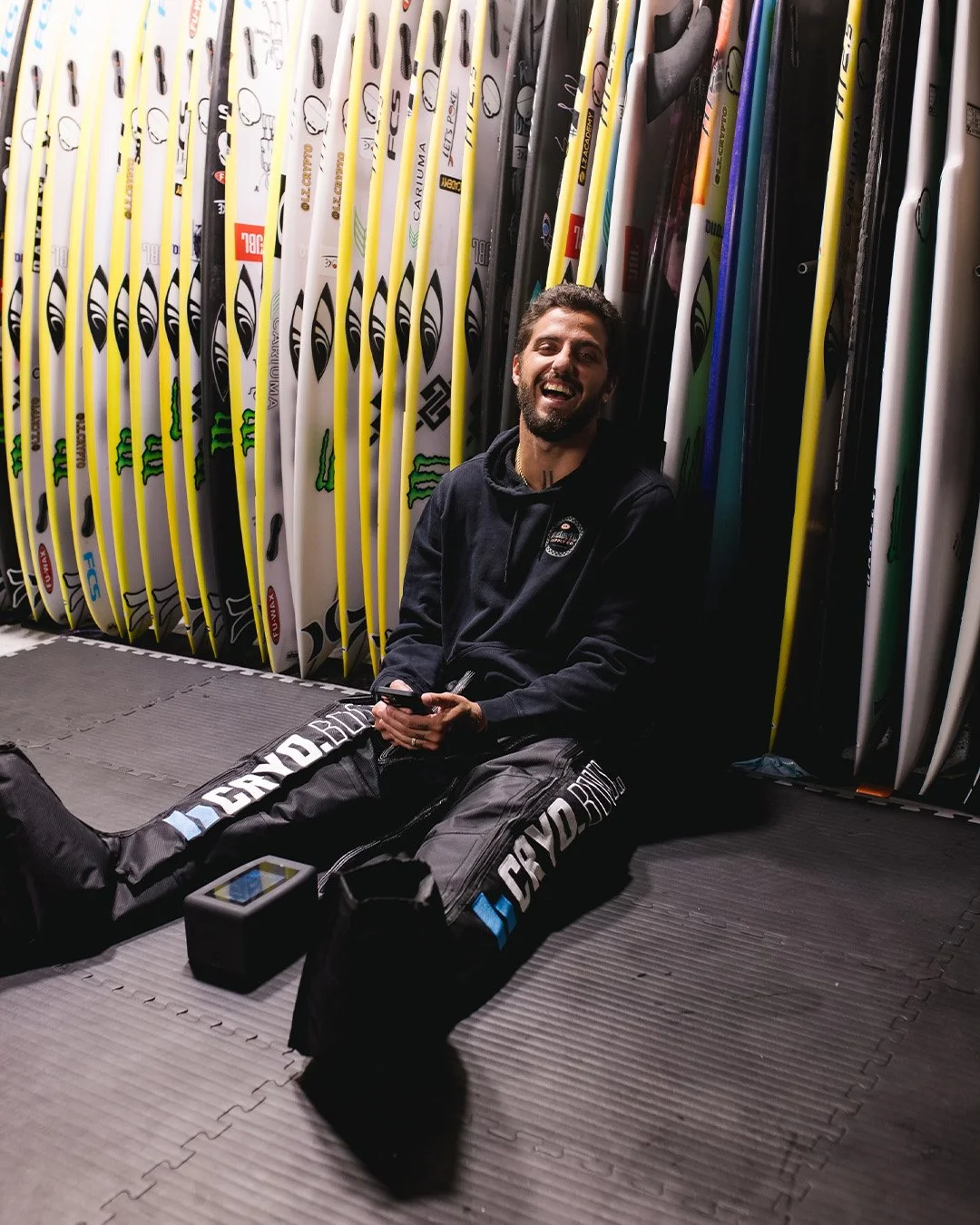 Hombre sonriente sentado en el suelo en una tienda de tablas de surf, rodeado de tablas de surf apiladas a su espalda.