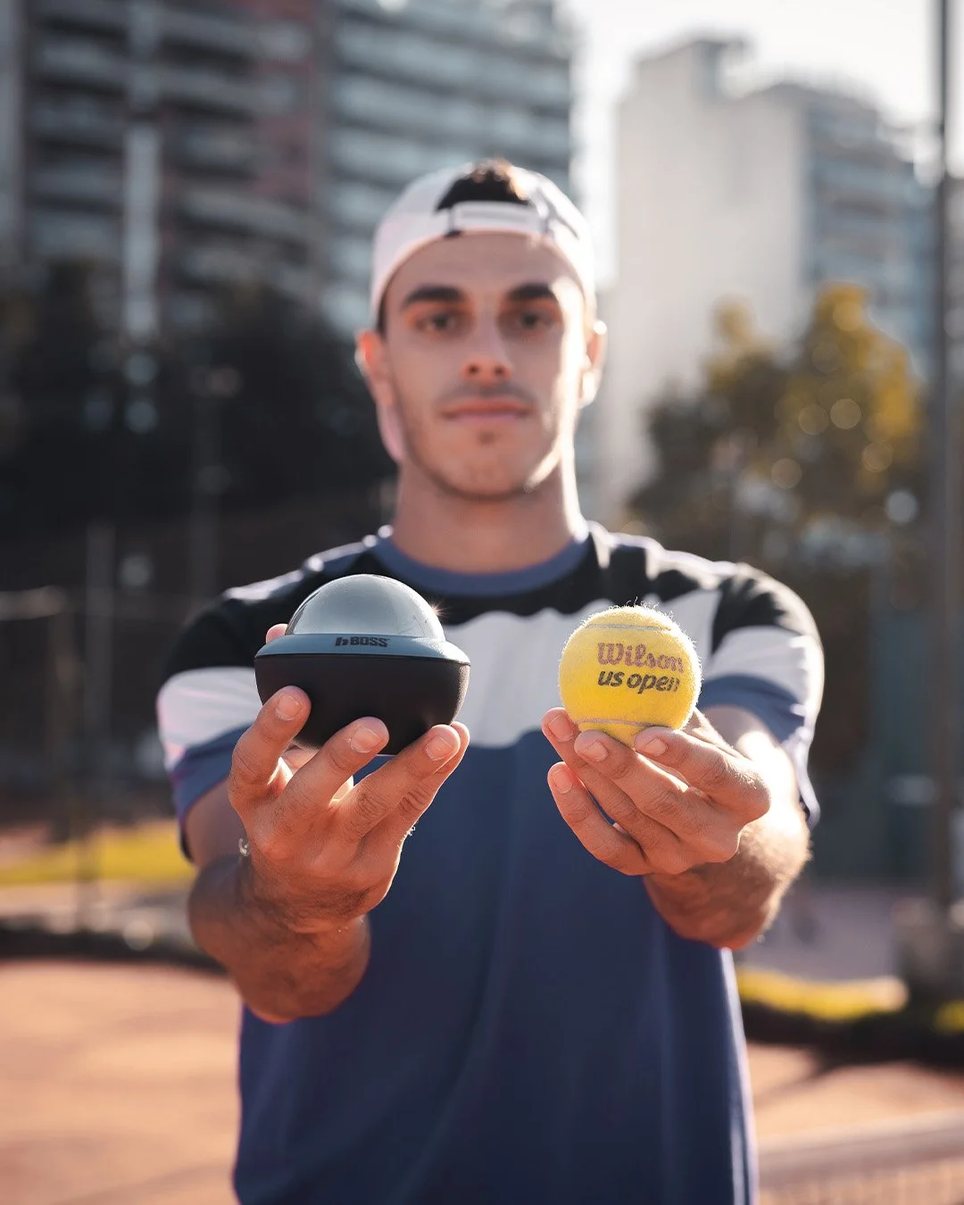 Joven con gorra, sosteniendo una pelota de tenis y un reloj de entrenamiento, en un parque urbano.