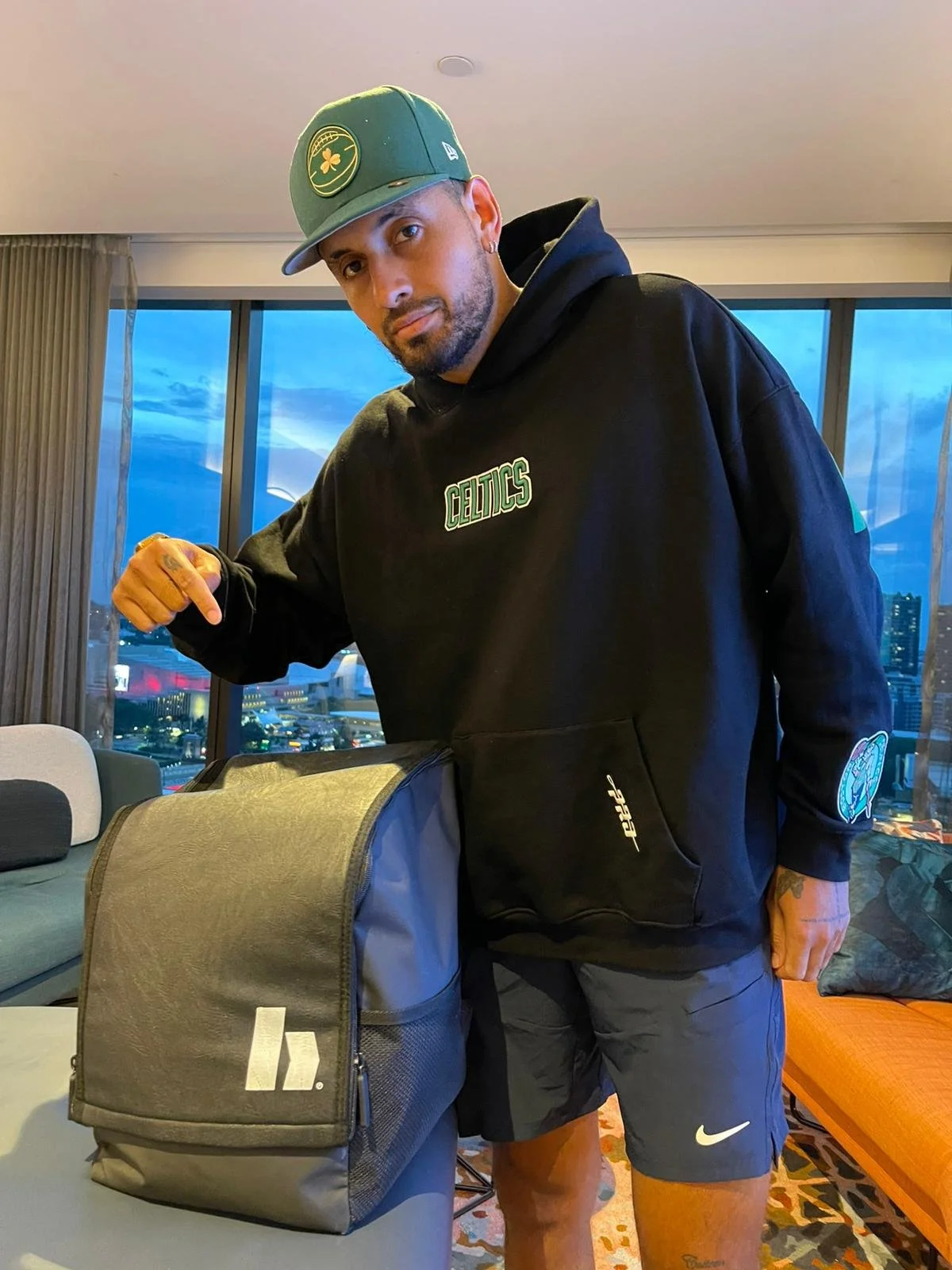 Hombre con gorra y sudadera de los Boston Celtics, haciendo gesto de fuerza con el puño, en una habitación con ventanas grandes y vista urbana durante la noche.