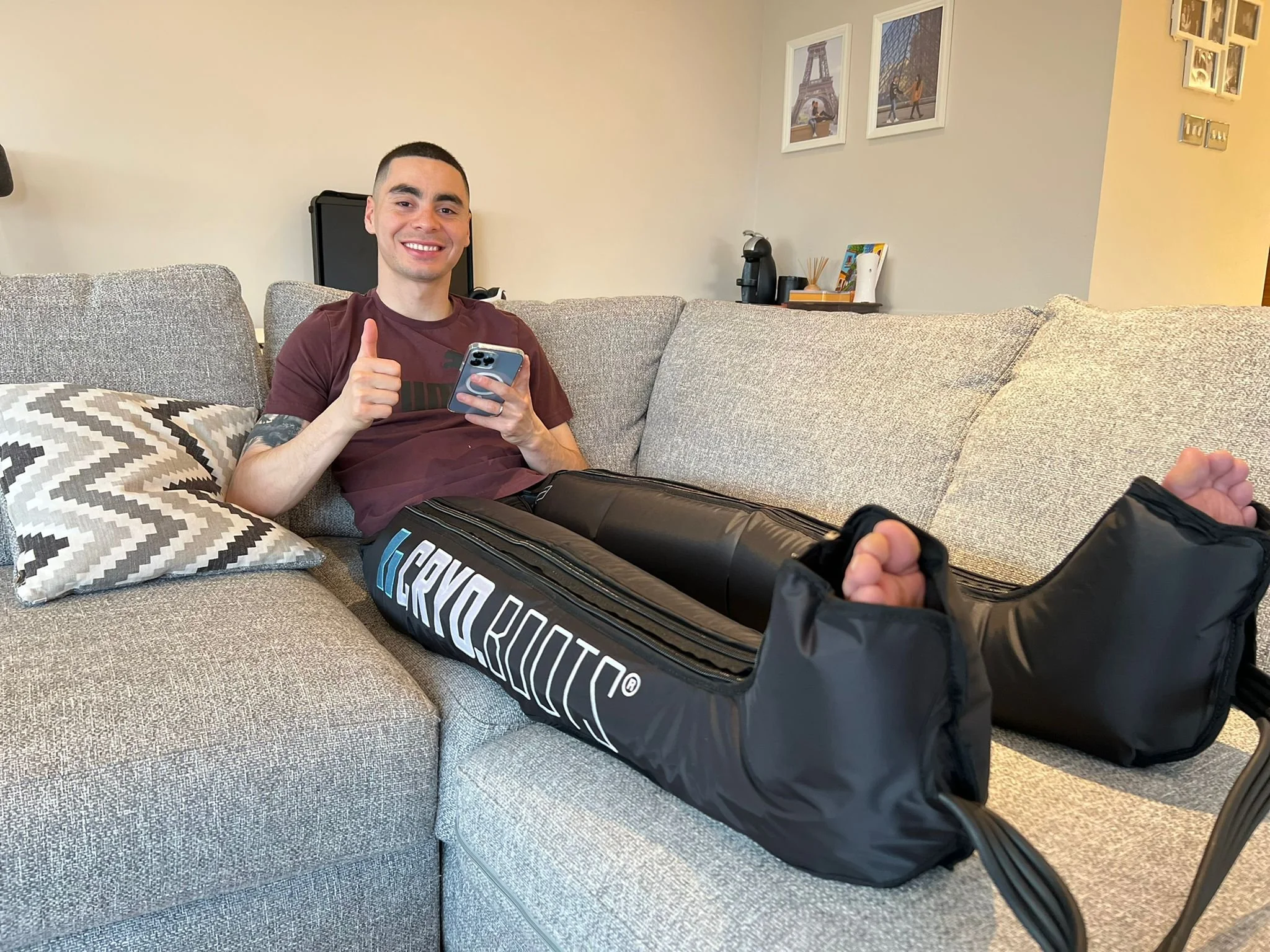 Joven sonriendo en un sofá con un teléfono móvil en la mano y equipo de golf en la pierna, en una sala con cuadros en la pared y objetos decorativos en una mesa.