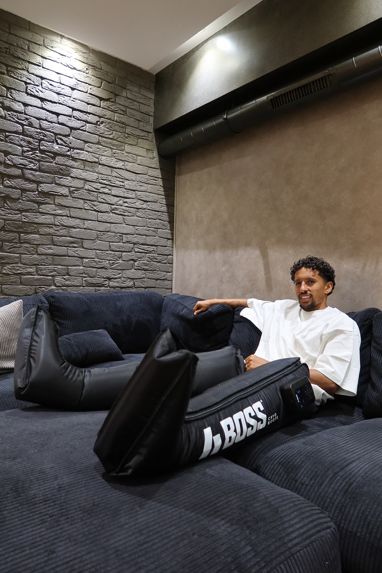 Hombre sentado en un sofá oscuro con varias almohadas, usa ropa blanca, en un ambiente con paredes de ladrillo y decoración moderna.