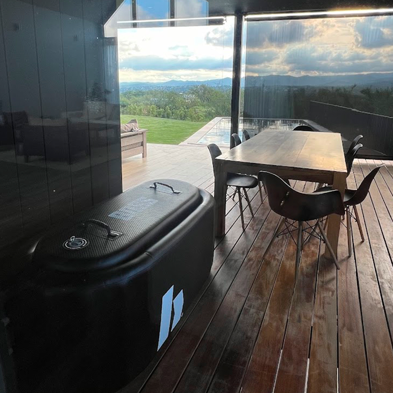Sala de comedor con mesa de madera y sillas negras, vistas a un paisaje natural con montañas en el fondo, en una terraza de madera con gran ventana de cristal.