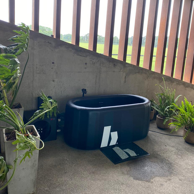 Bañera de hidroterapia negra en un espacio con plantas y vista al campo desde un balcón con barrotes de madera.