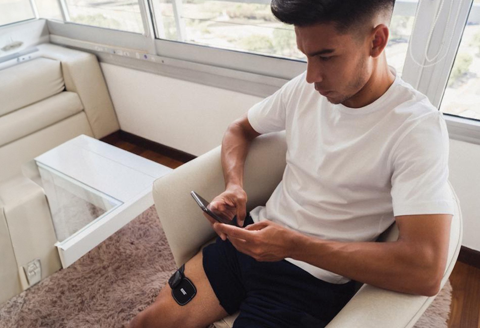Joven sentado en un sofá de color crema, usando una camiseta blanca y pantalones oscuros, con un monitor de frecuencia cardíaca en la pierna izquierda, en una habitación con ventanas grandes y cortinas. Está mirando su teléfono móvil.