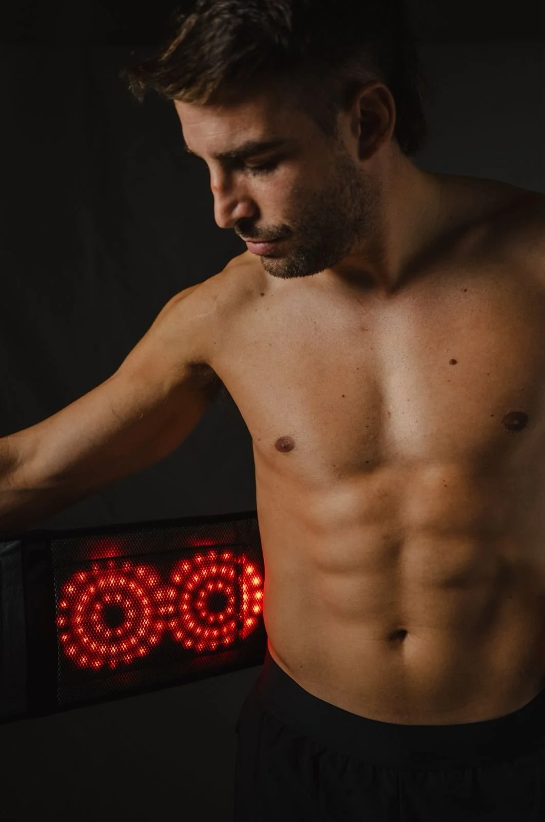 Hombre sin camiseta en primer plano con un fondo negro y una señal de luces rojas en forma de círculo.