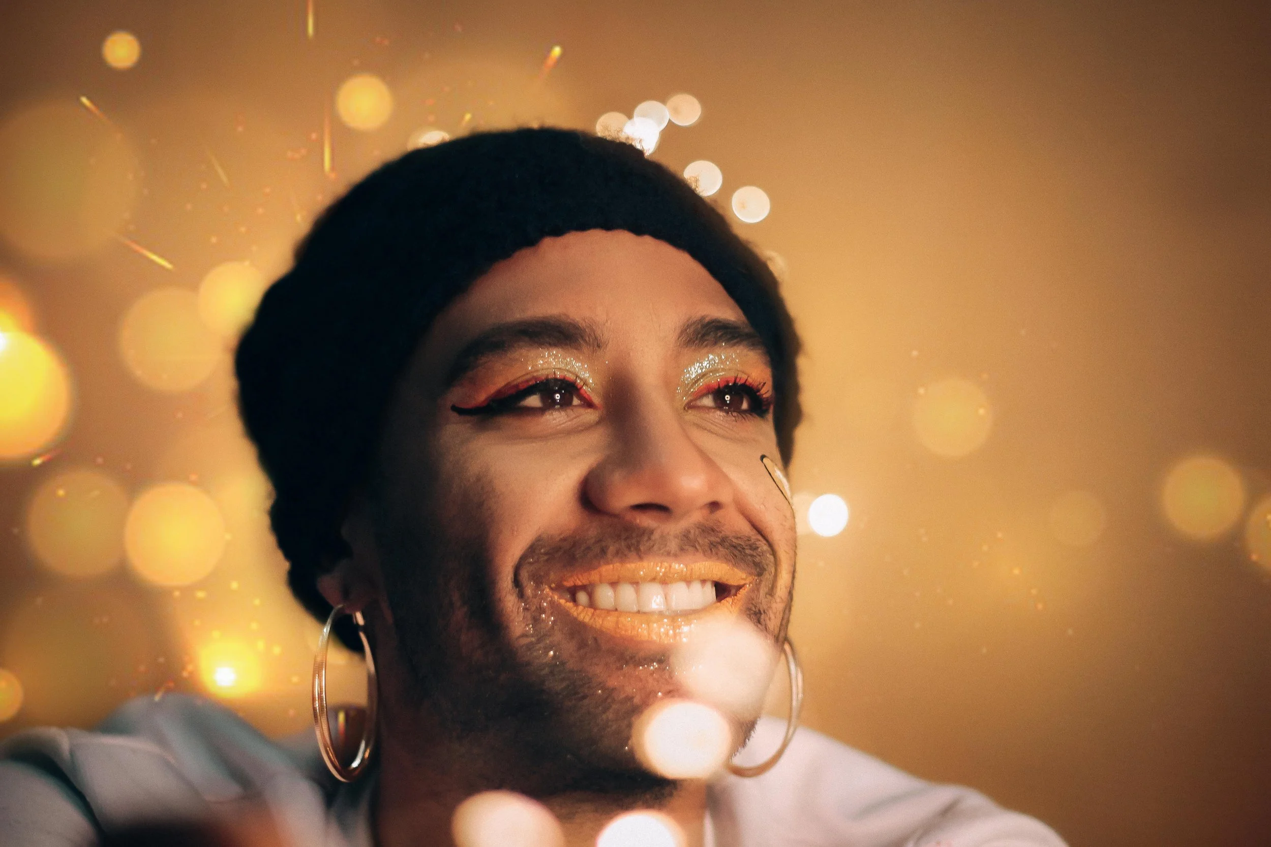 An 2SLGBTQIA+ person with glitter on their face smiles.