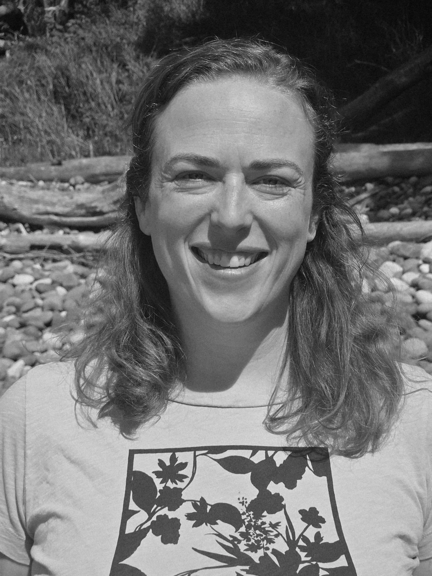 Black and white photo of a smiling woman with shoulder length brown hair standing in front of driftwood.
