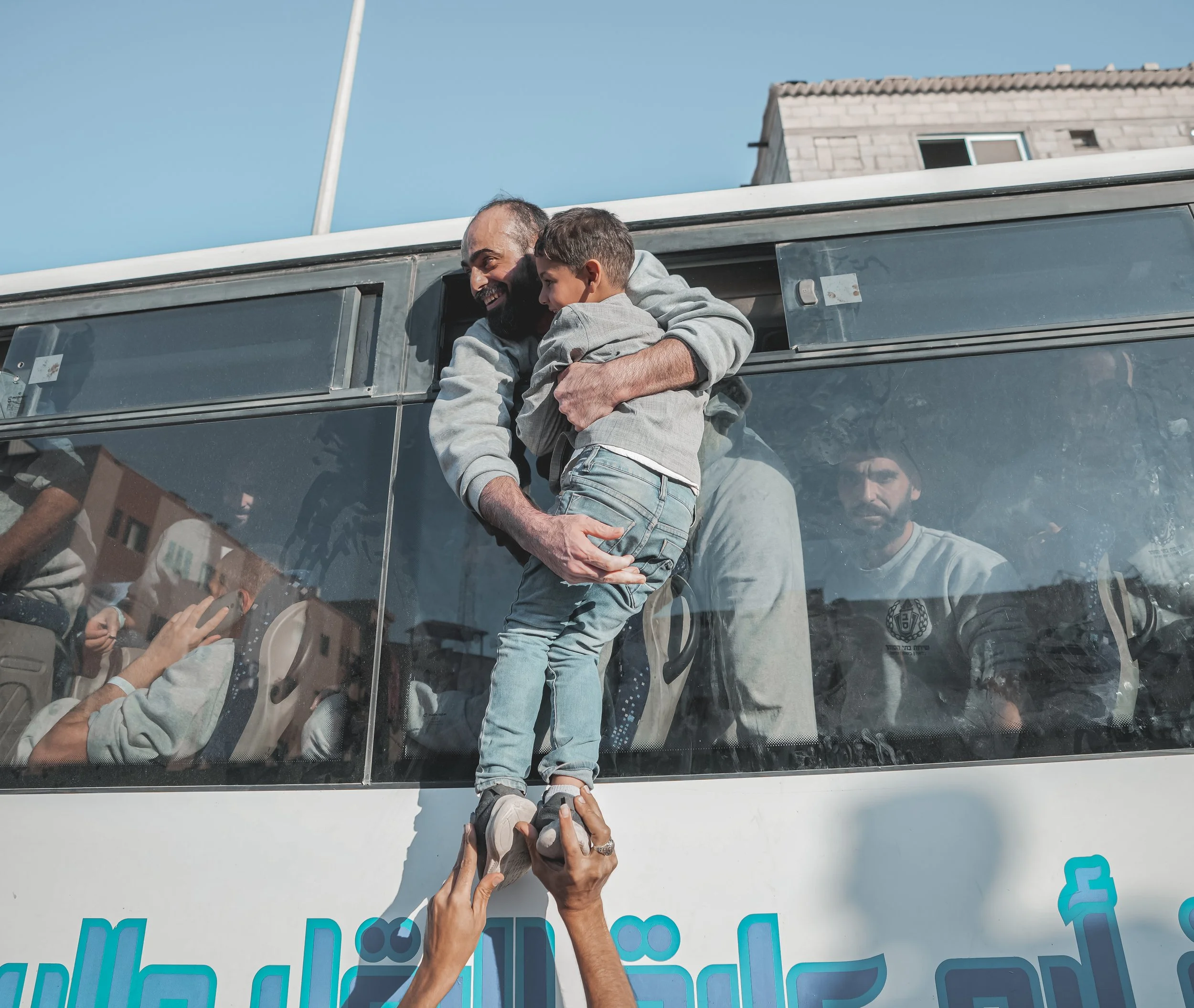 A Palestinian prisoner, standing inside a bus full of man, embraces a young boy through the vehicle window. The young boy is outside the bus and held up by his feet to the window.