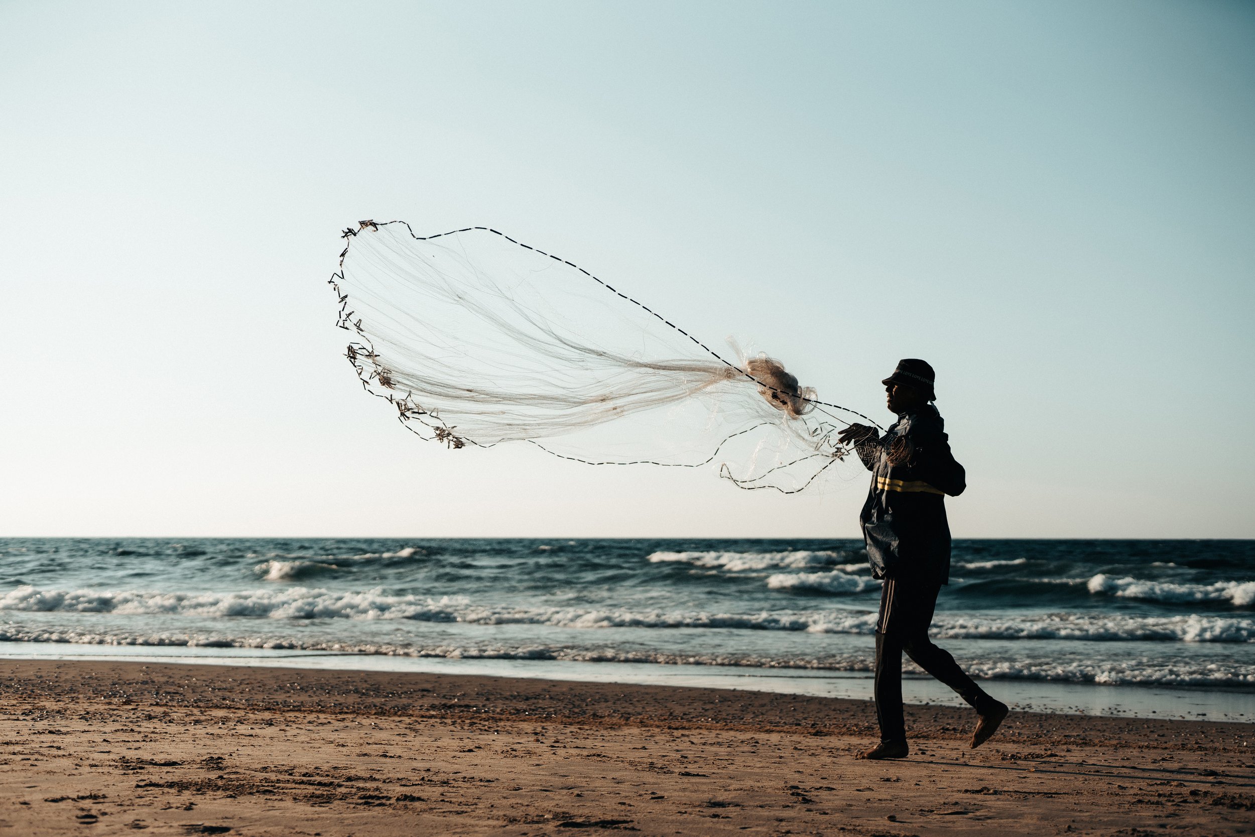 A person standing on a sandy beach, casting a fishing net into the ocean during sunset.
