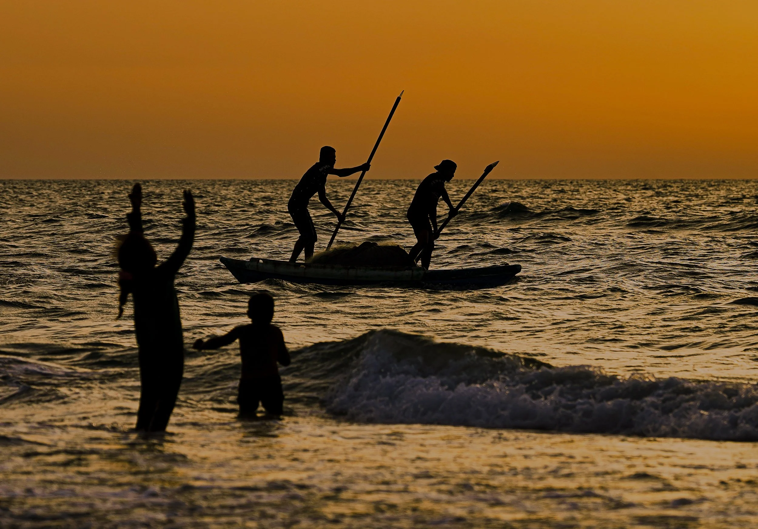 Silhouettes of two Palestinians paddling on a boat, fishing in the water during sunset, with others in the water nearby, and a warm orange sky in the background.