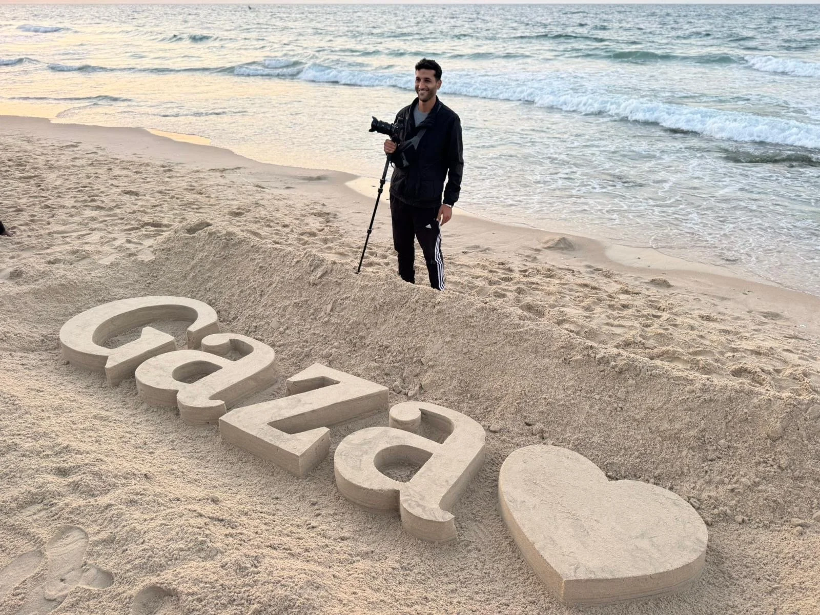 A Palestinian photographer with a camera on a tripod stands on the beach in Gaza, near a large sand sculpture spelling out 'Gaza' with a heart. The ocean waves are in the background.