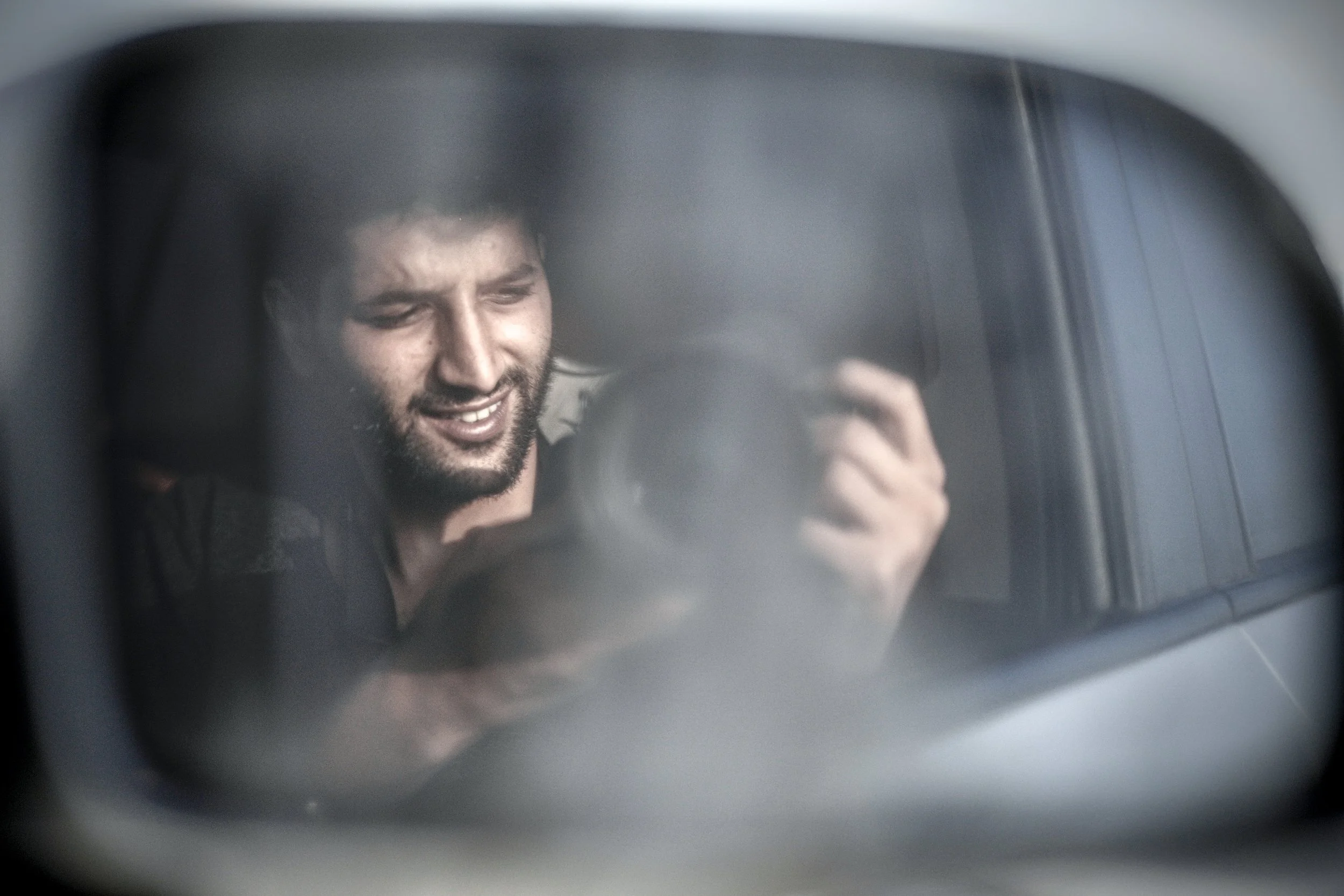 A man smiling and looking at his camera, reflected on a car's side-view mirror.