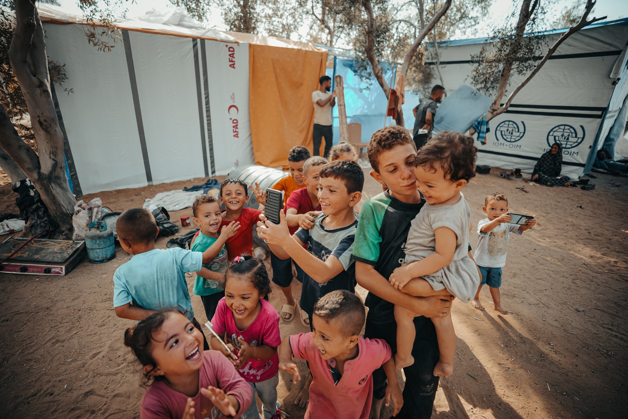 A group of Palestinian children playing and smiling outdoors in a sandy area with tents and trees in the background.