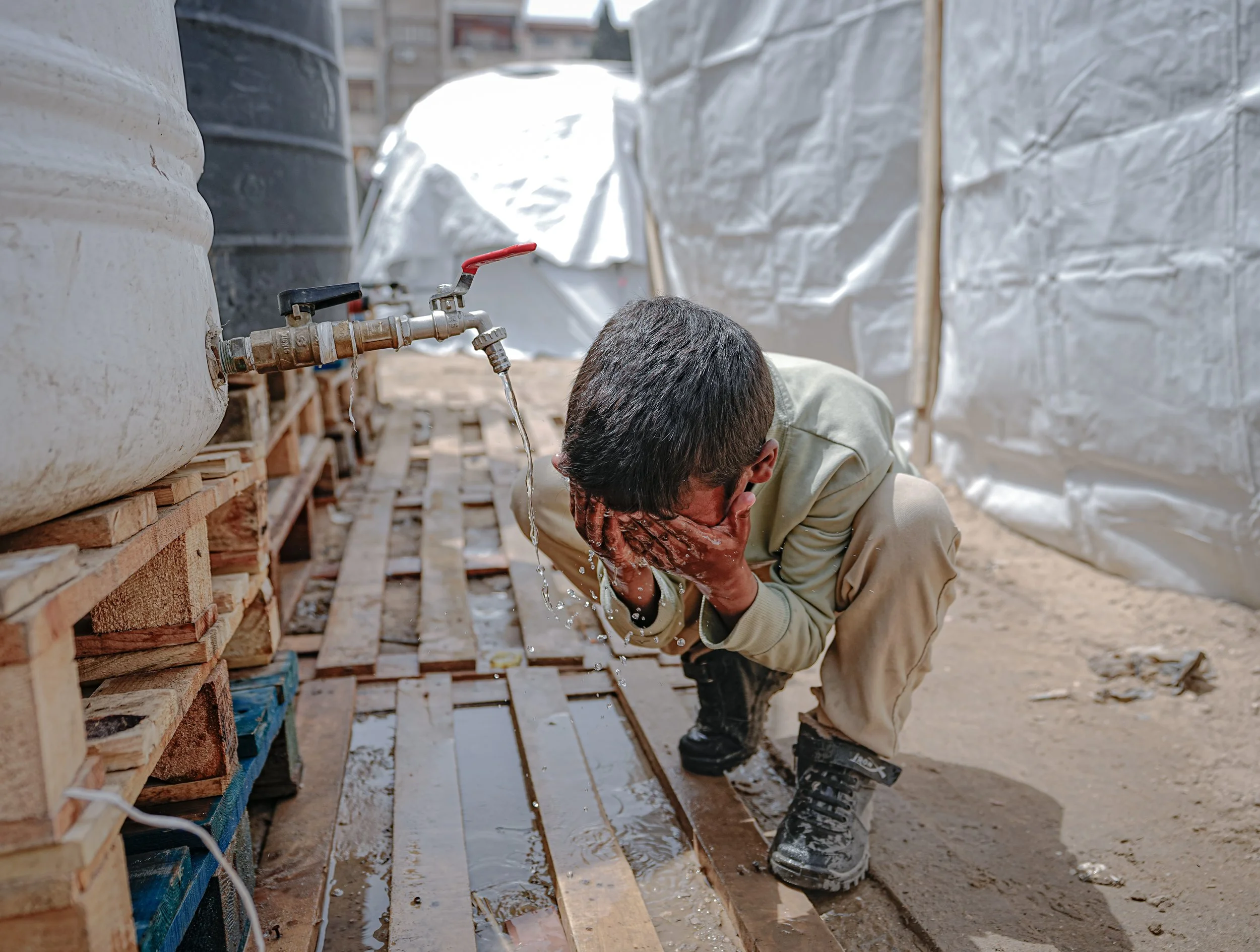 A young Palestinian boy kneeling down in a displacement tent area, drinking water from his hands under a water faucet.