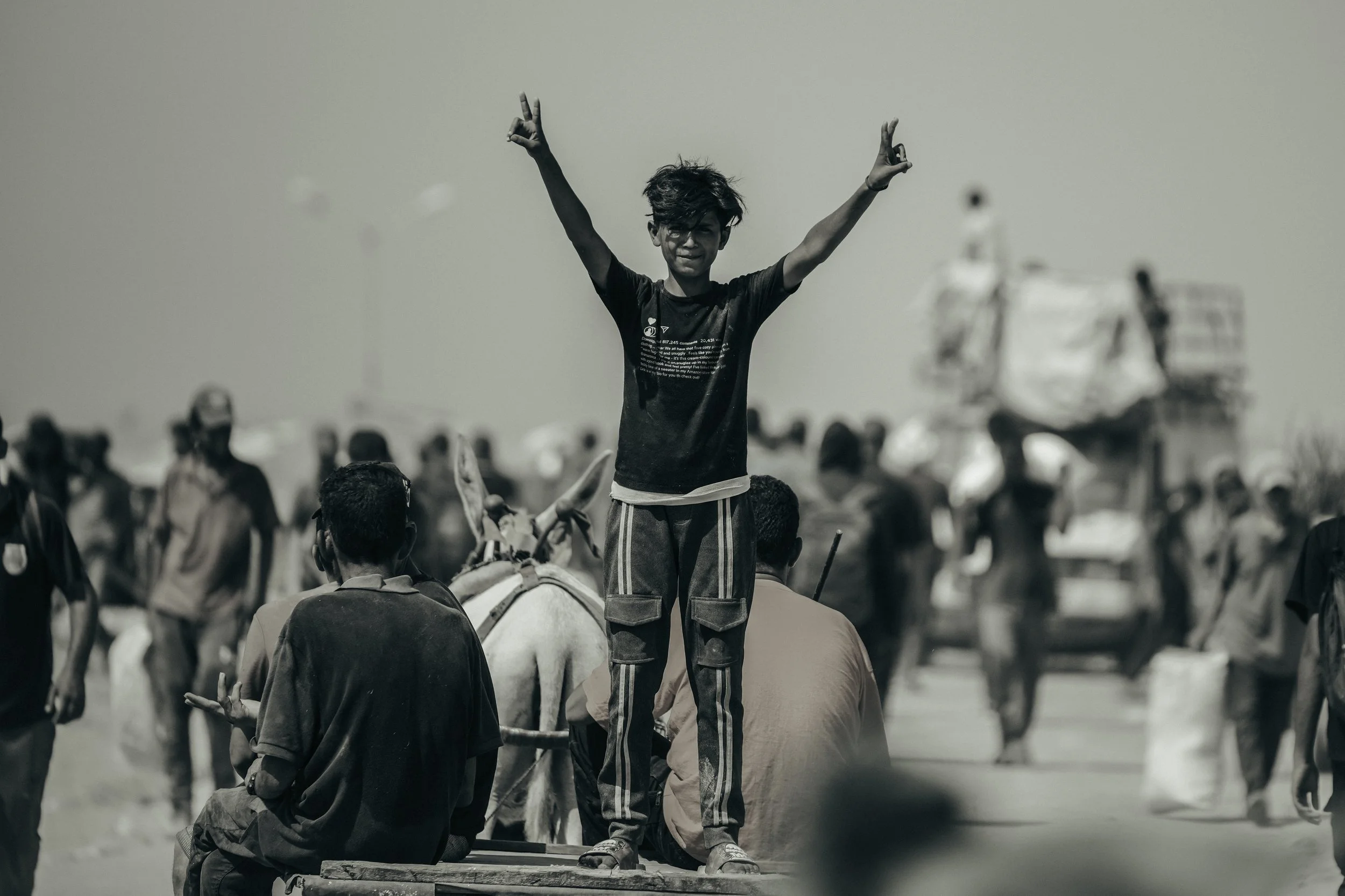 A young Palestinian boy standing on a vehicle with his arms raised and making a peace sign with both hands, surrounded by a crowd of people, in a black and white photo, in Gaza.