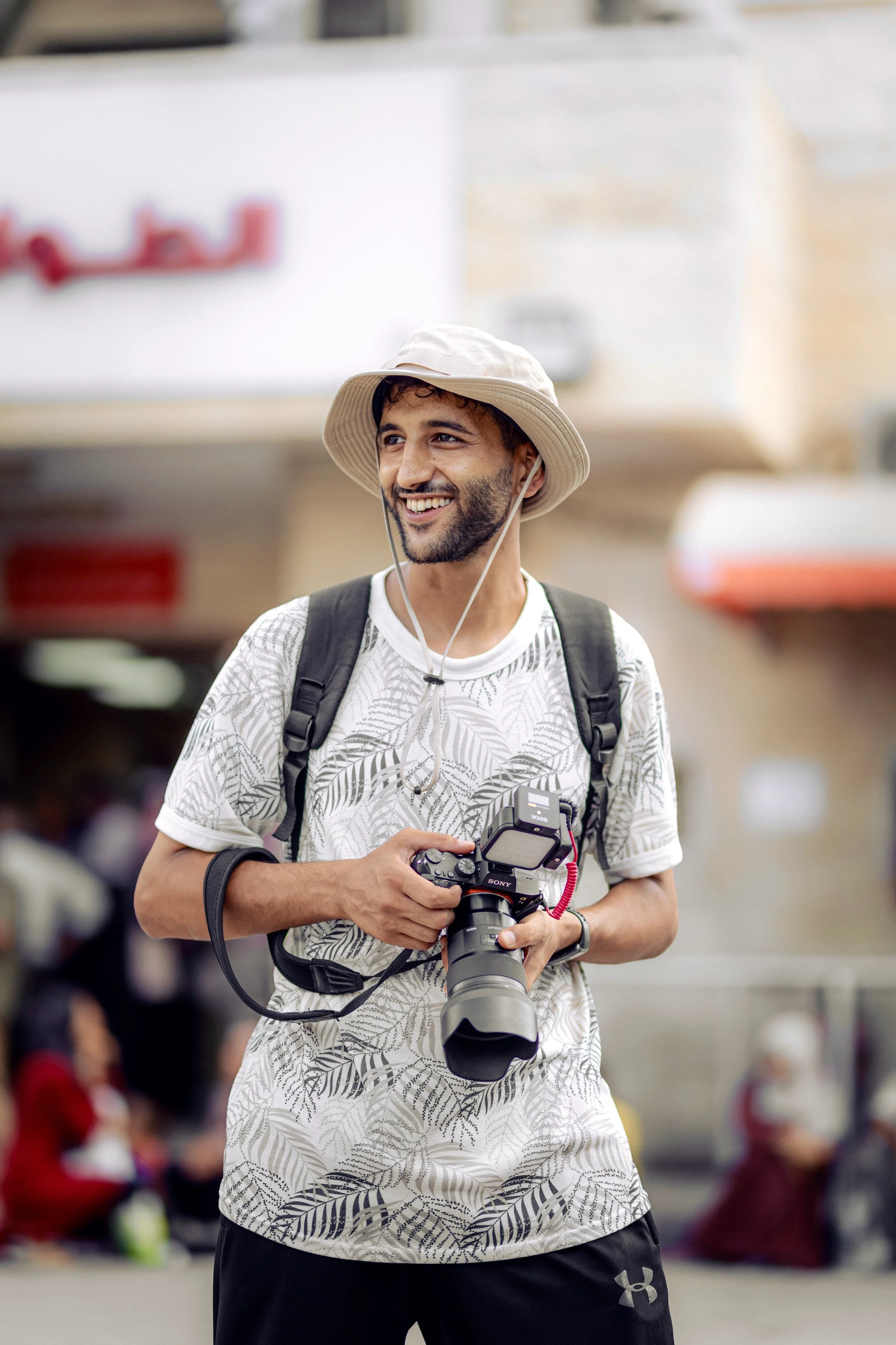 A Palestinian photographer smiling while holding a professional camera in front of a hospital in Gaza.