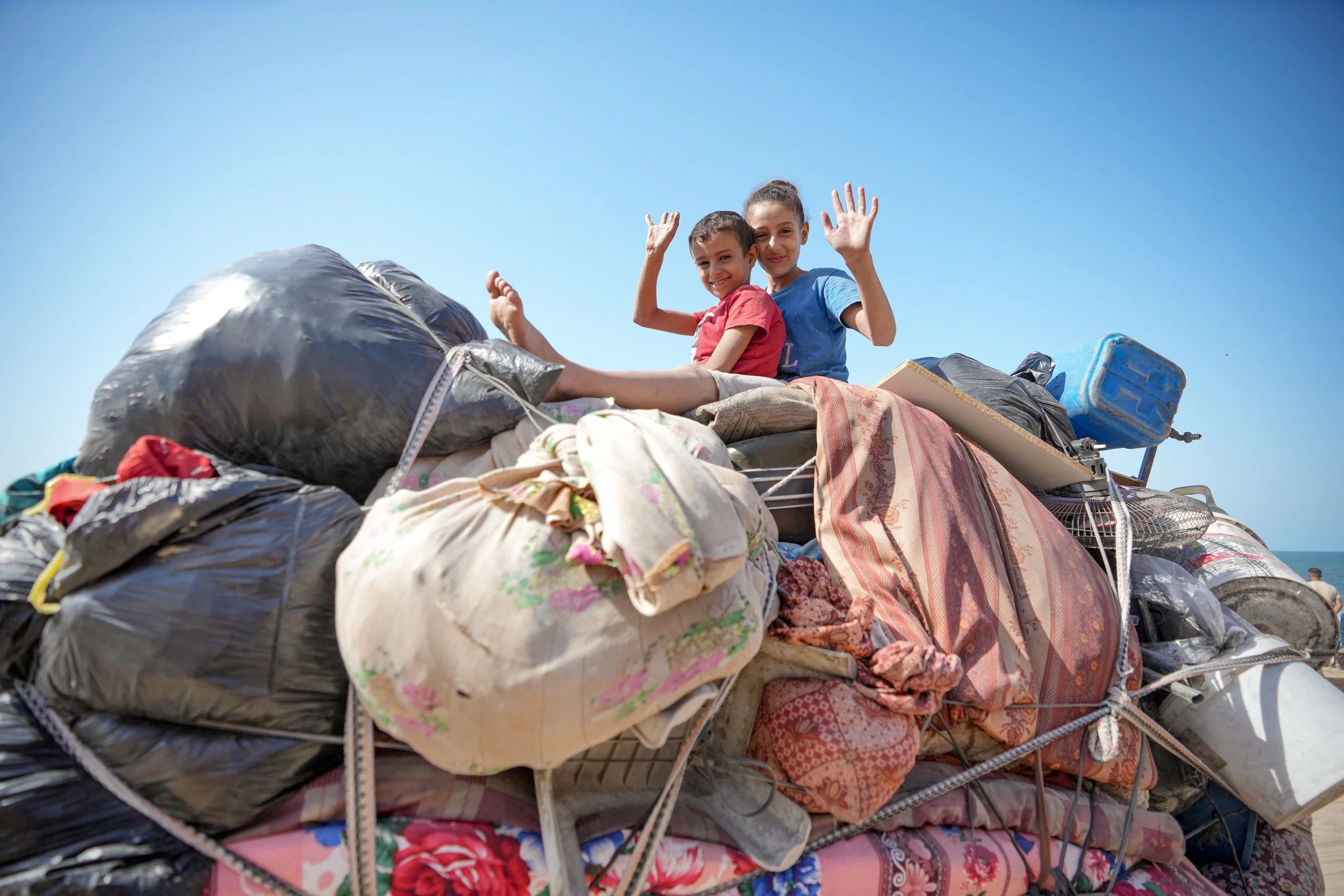 Two Palestinian children smiling and waving while sitting on a large pile of bags and belongings, under a clear blue sky, during a displacement.