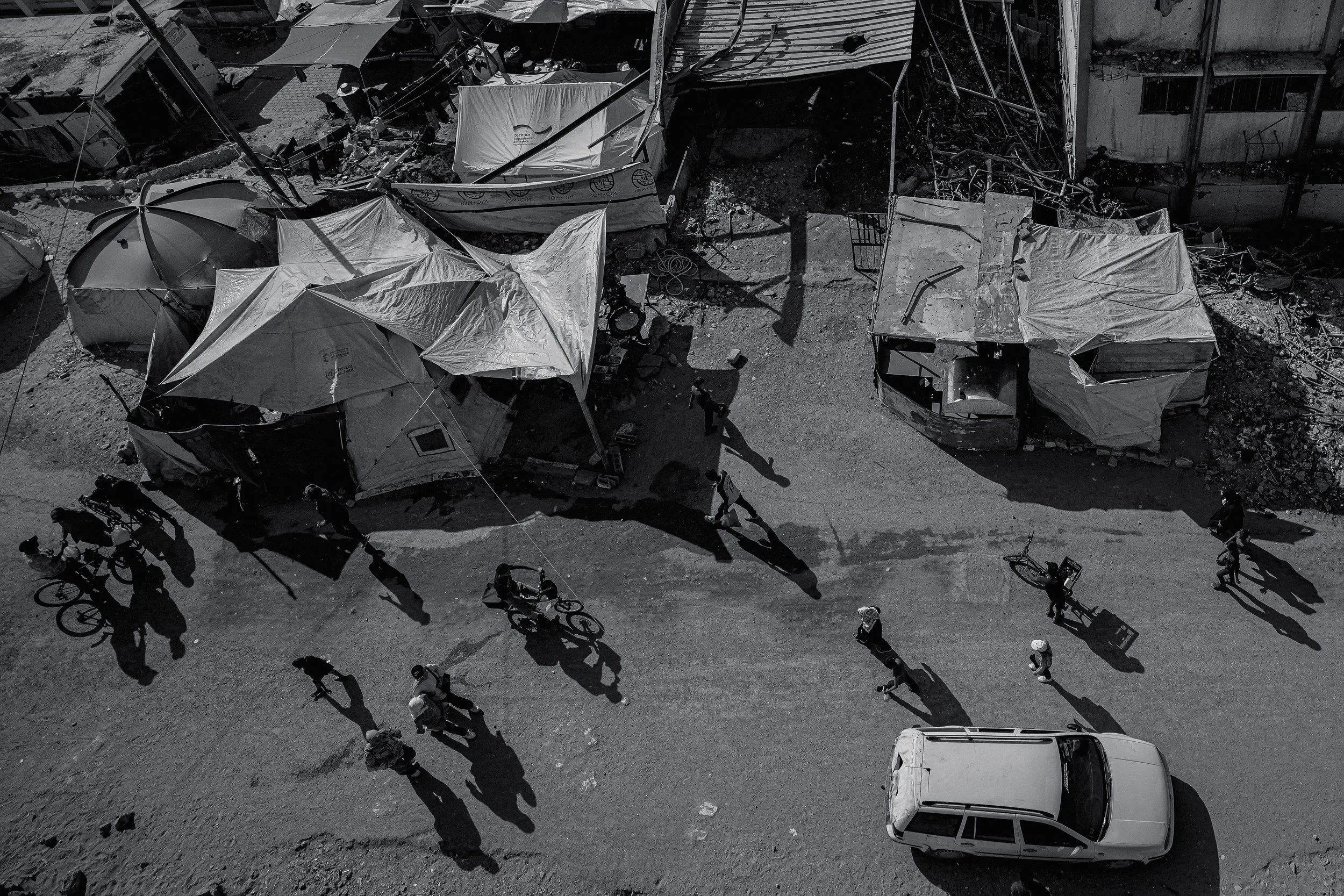Black and white aerial photo of a street scene showing a damaged bus and tents, with people and bicycles walking nearby, in Gaza.
