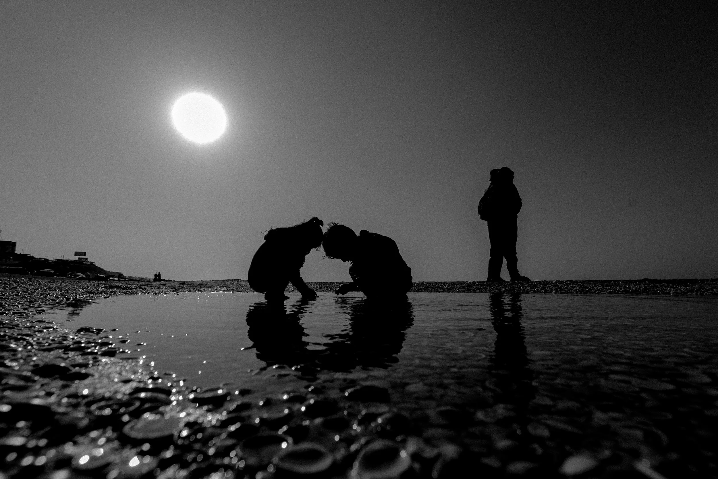 Silhouette of four children at the beach in Gaza during sunset, with three crouching by a shallow pool and one standing nearby, all reflected in the water, under a bright sun in the sky.