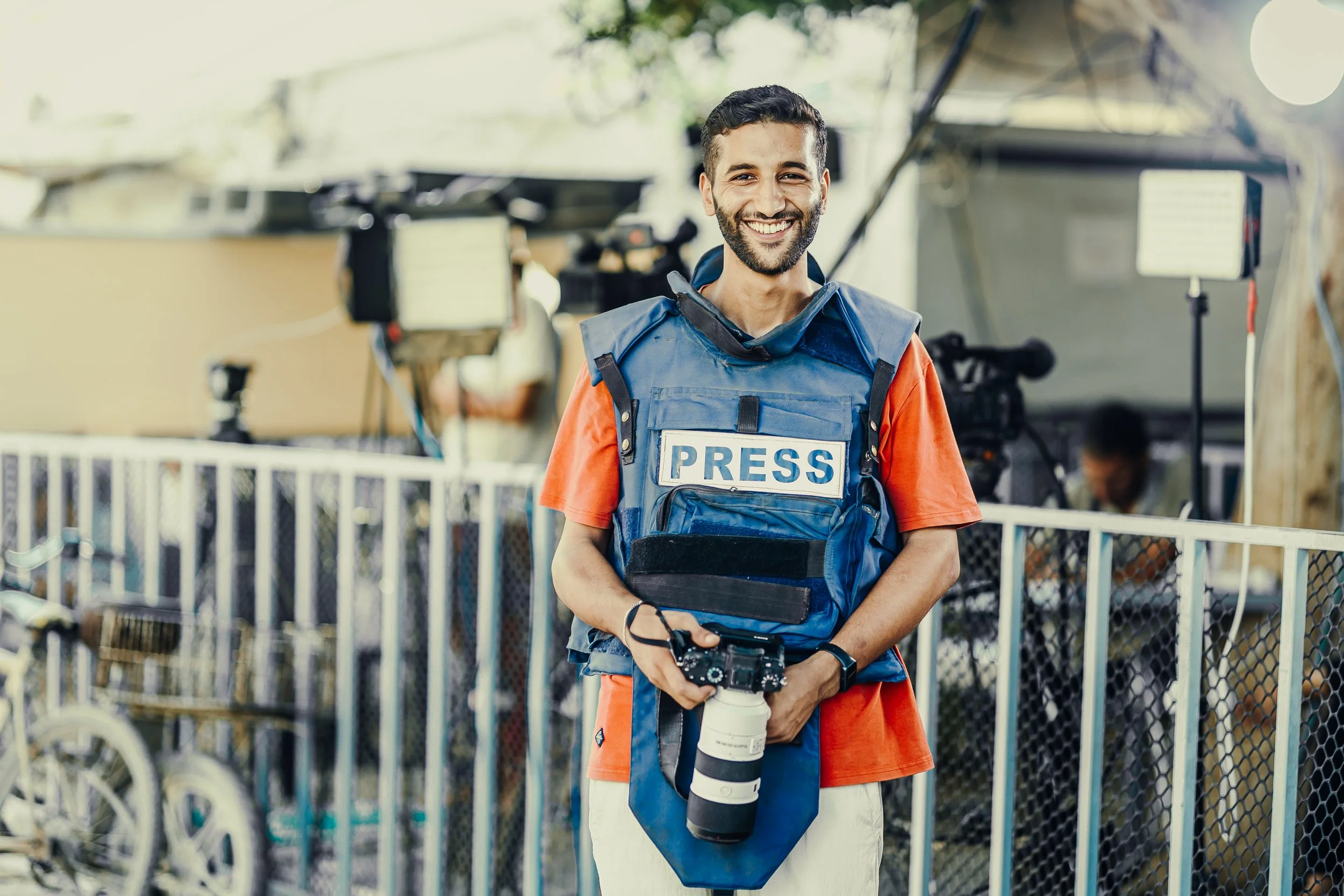 A smiling male photographer wearing a press vest and holding a camera with a telephoto lens stands in a press work area in Gaza.
