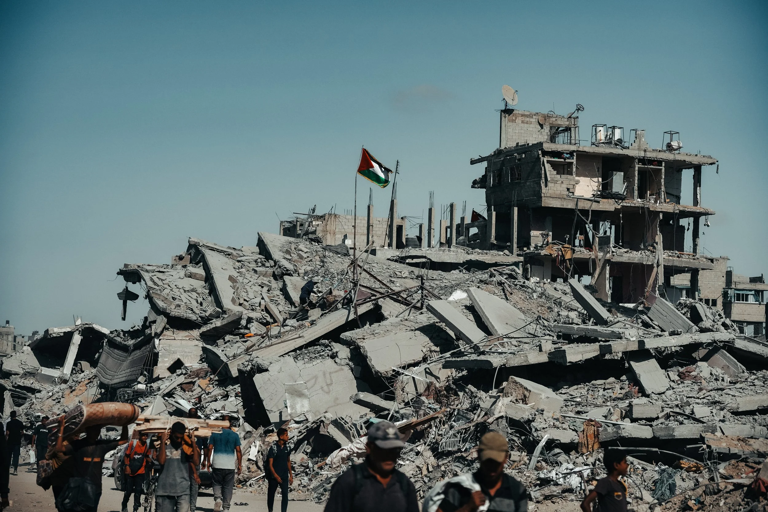 People walking through the debris of a destroyed building with collapsed concrete and rubble, and a Palestinian flag seen standing upright amidst the destruction.
