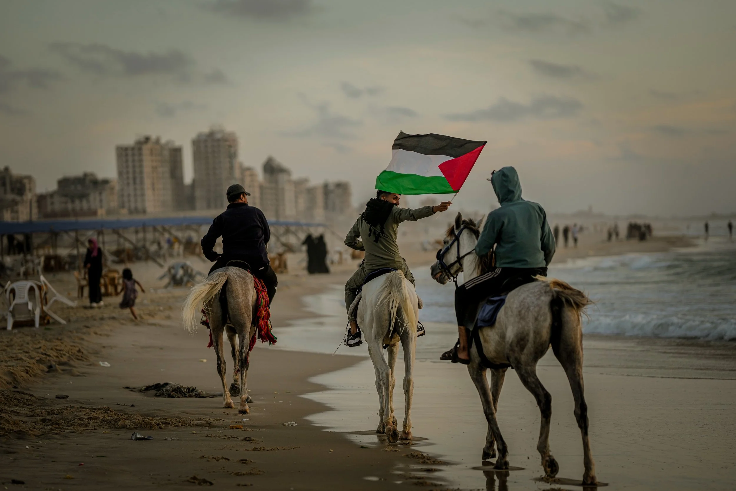 Palestinians riding horses along the beach in Gaza, with one individual holding a Palestinian flag. Buildings are visible in the distance under a cloudy sky.