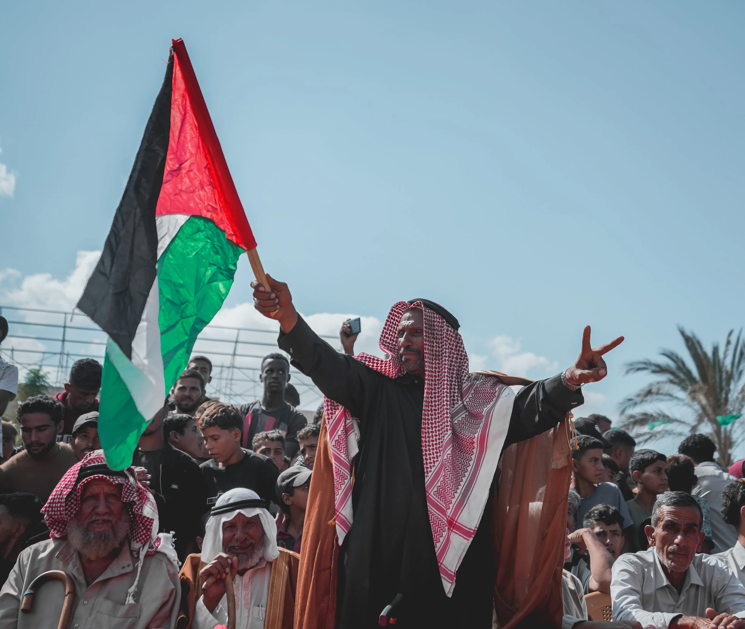 A large crowd of Palestinian men with gathered outdoors under a blue sky, some wearing traditional Palestinian attire. One man in the center, wearing a keffiyeh, is holding a Palestinian flag and making a peace sign with his hand. 
