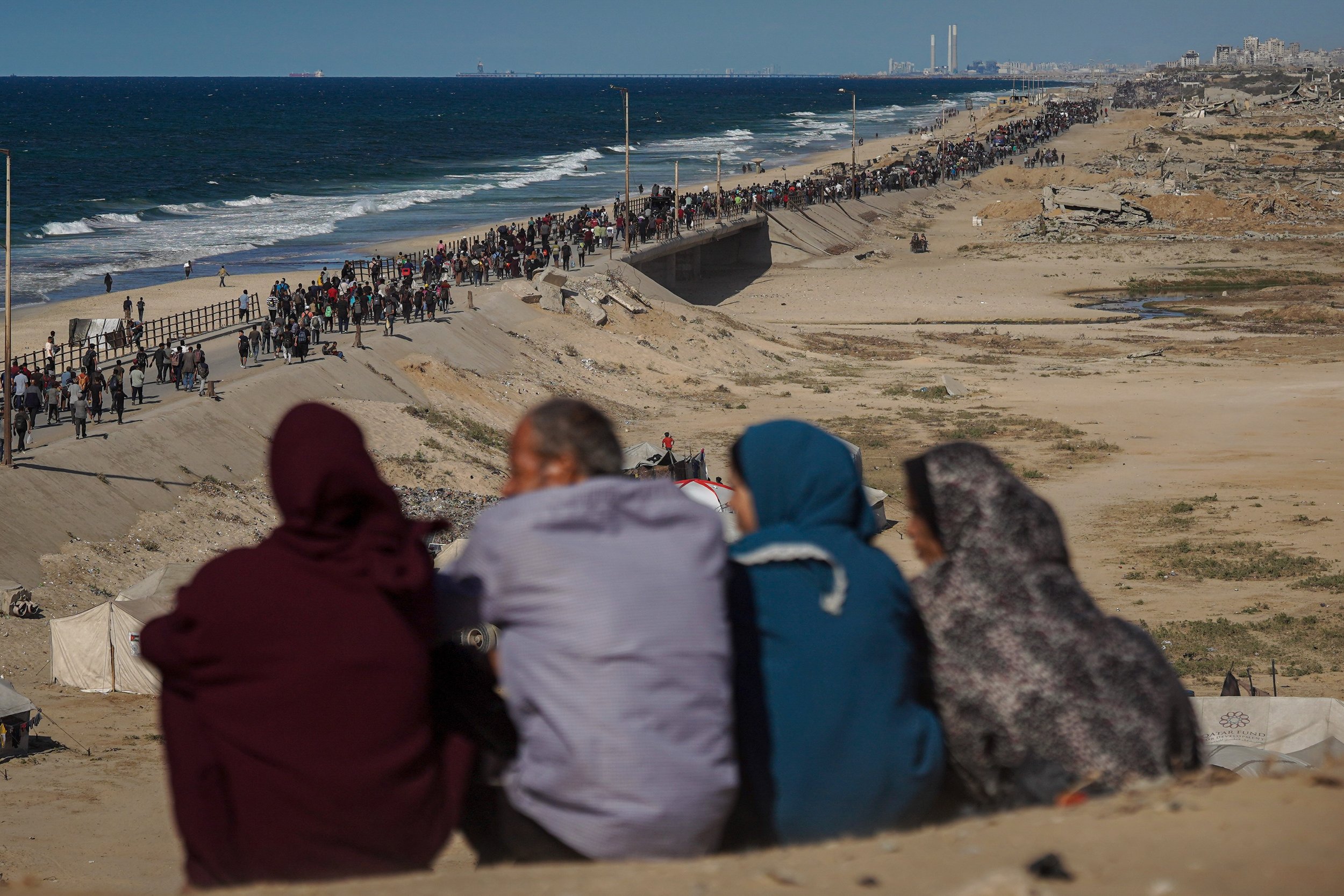 Four Palestinian adults watch a long queue of Palestinians walking along the beach road in Gaza during a displacement.