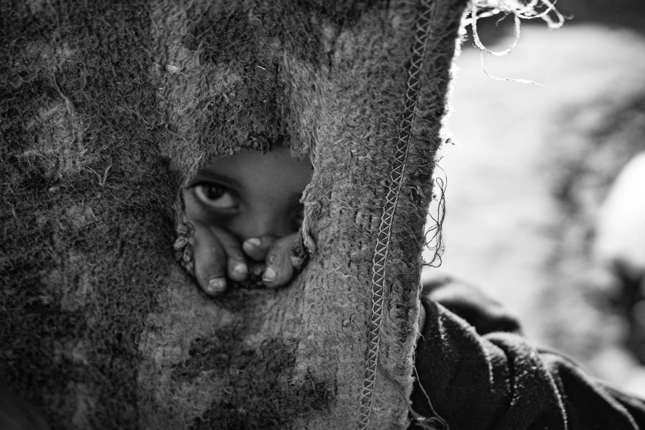 A young Palestinian child peering through a small opening in a torn piece of rough fabric from a tent.