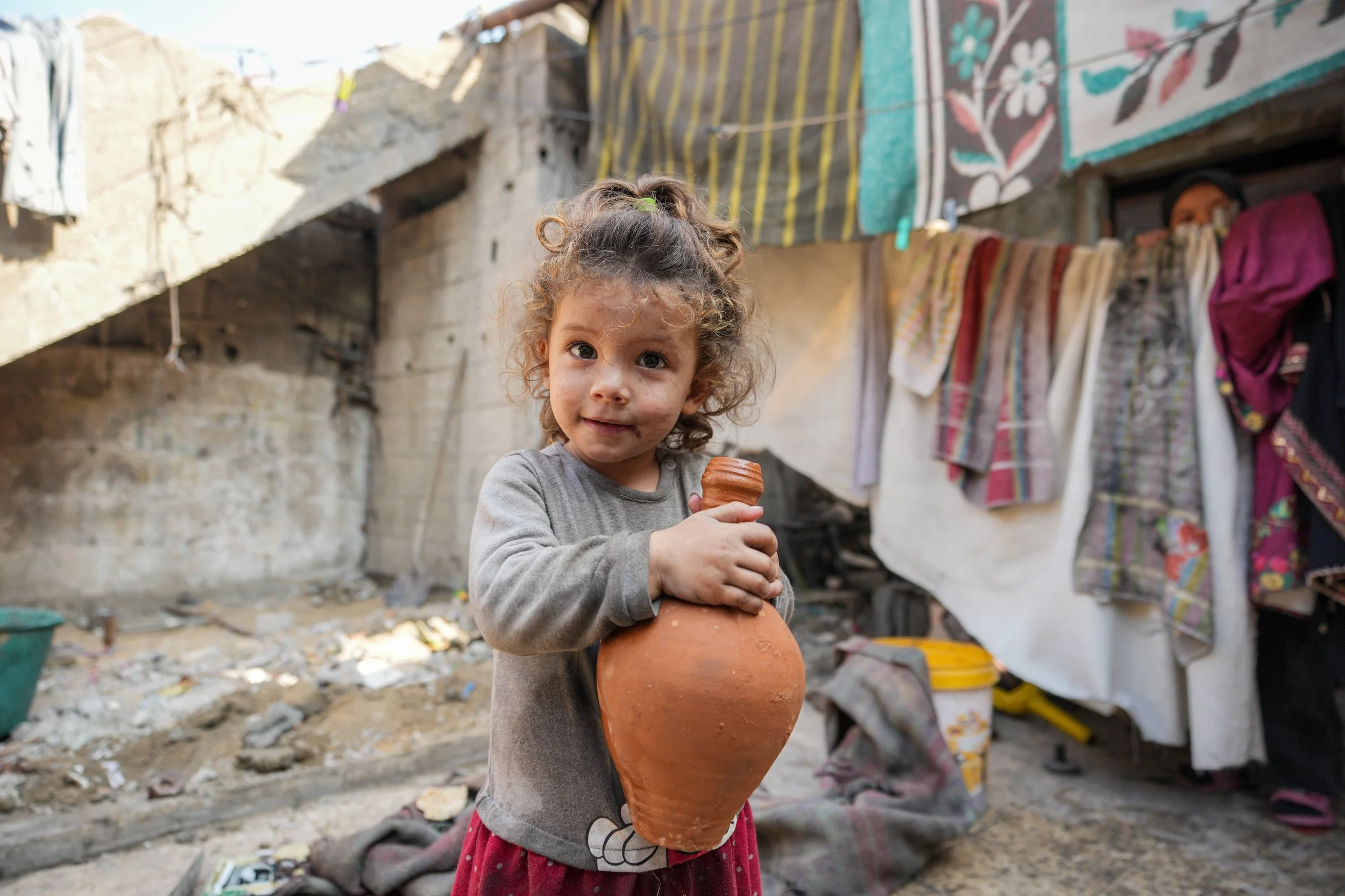 Young Palestinian girl with curly hair holding a large clay pot outside a tent area.